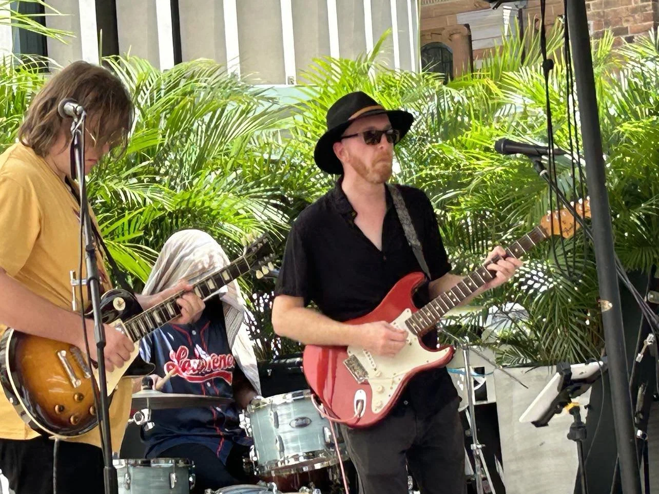 Three musicians playing instruments outdoors with green plants behind them.