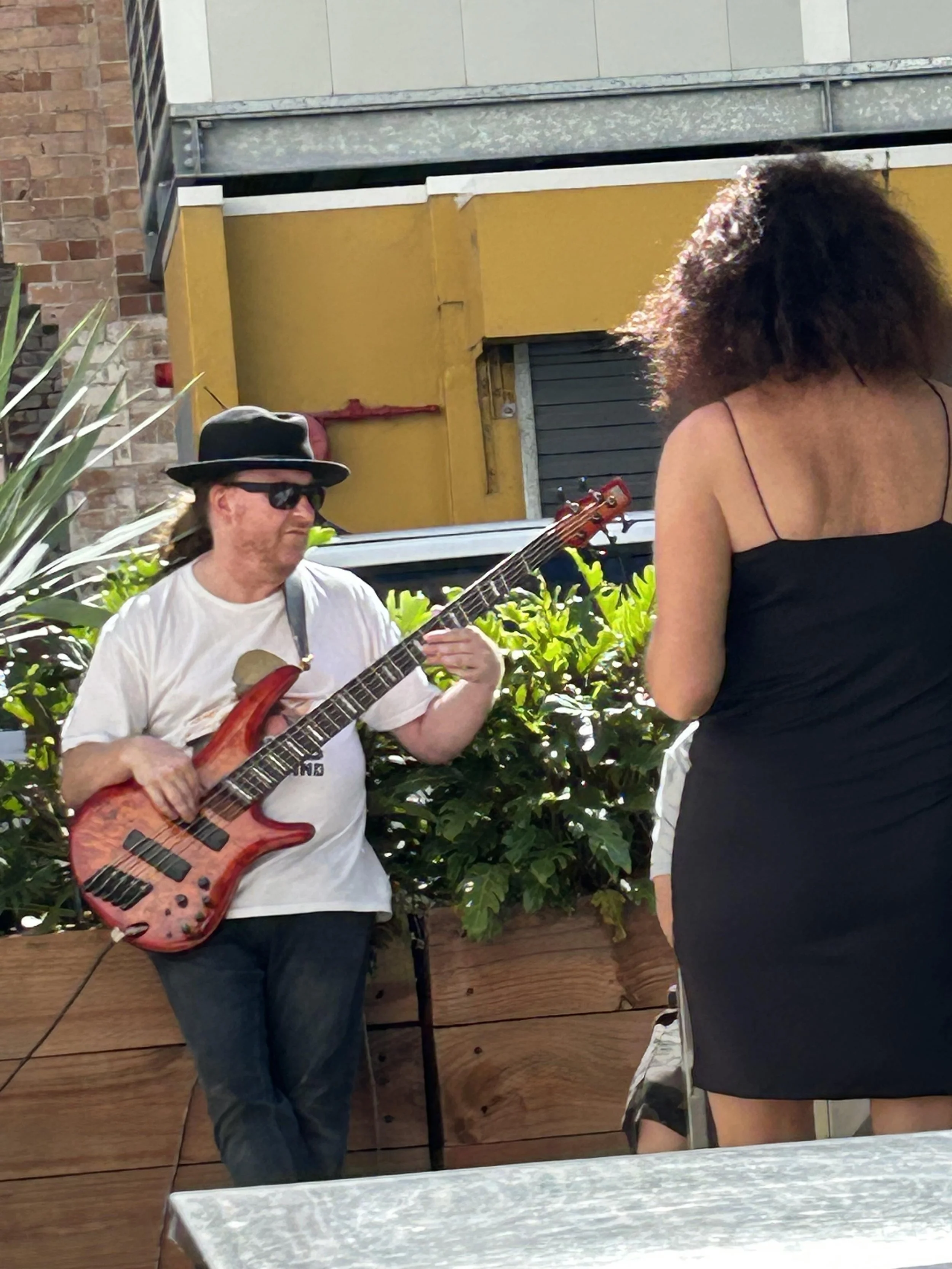 A man wearing sunglasses, a black hat, and a white T-shirt playing an electric guitar in an outdoor setting with a yellow wall, plants, and a woman in a black dress in the background.