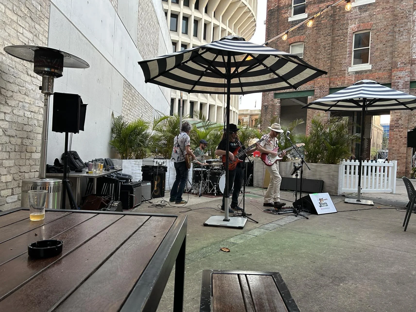 A band playing live music outdoors under large black and white striped umbrellas, with musicians playing guitars and drums. There are potted plants in the background and string lights above. The scene is set in an urban area with brick buildings.