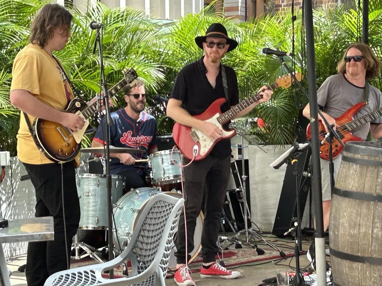 Group of four men playing musical instruments, with three of them playing guitars and one on drums, outdoors with green plants in the background.