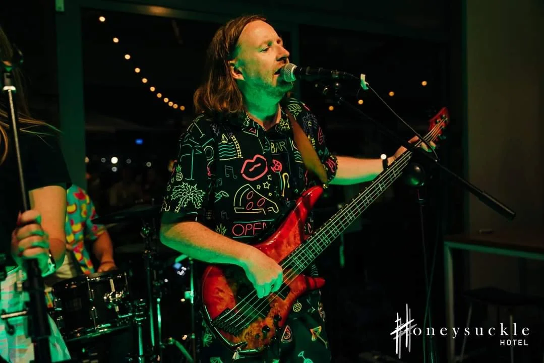 A man singing into a microphone while playing an electric guitar during a performance at a hotel, with dim lighting and string lights in the background.