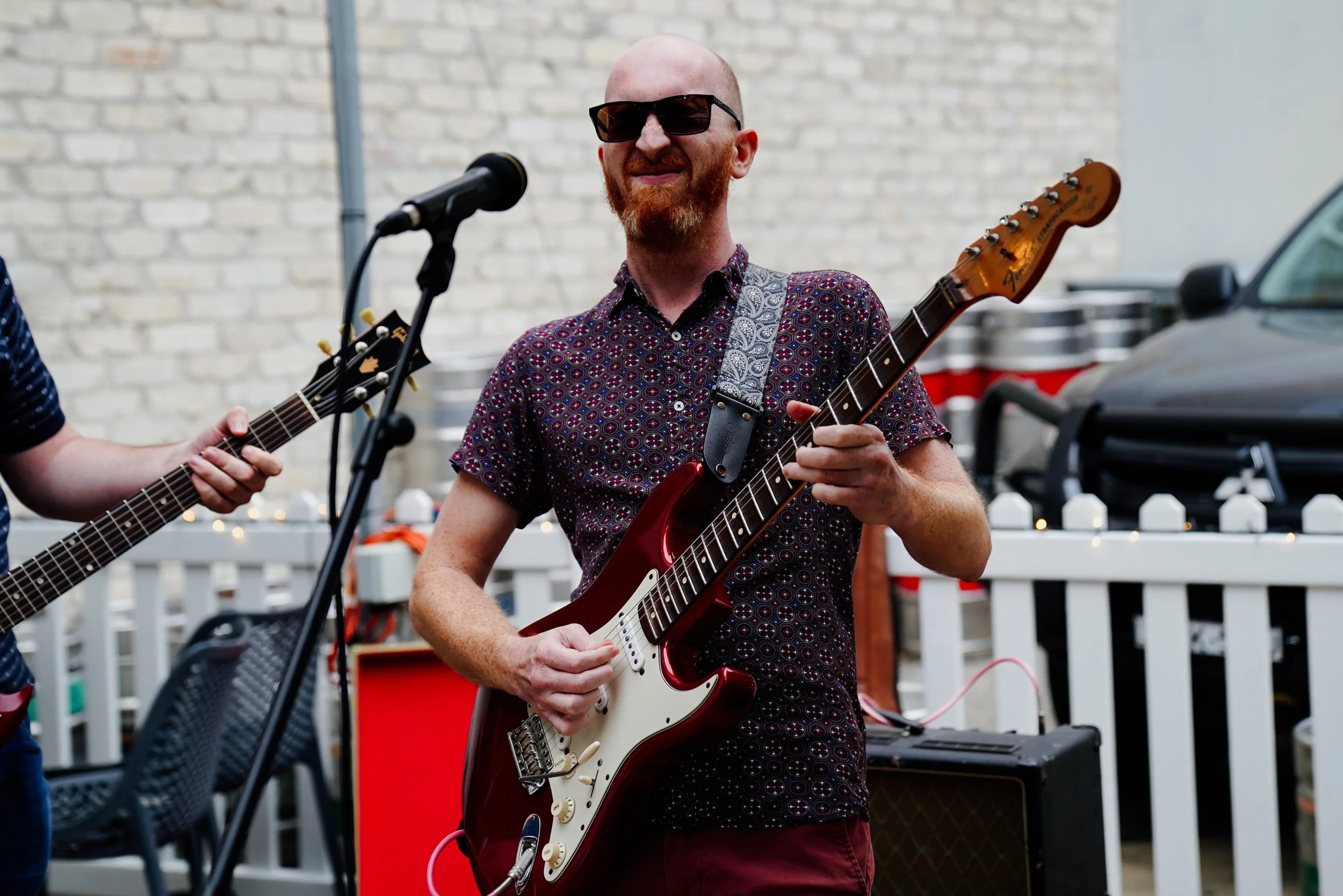 A man with a beard, sunglasses, and a patterned shirt is playing an electric guitar at an outdoor event, near a microphone with a brick wall in the background.
