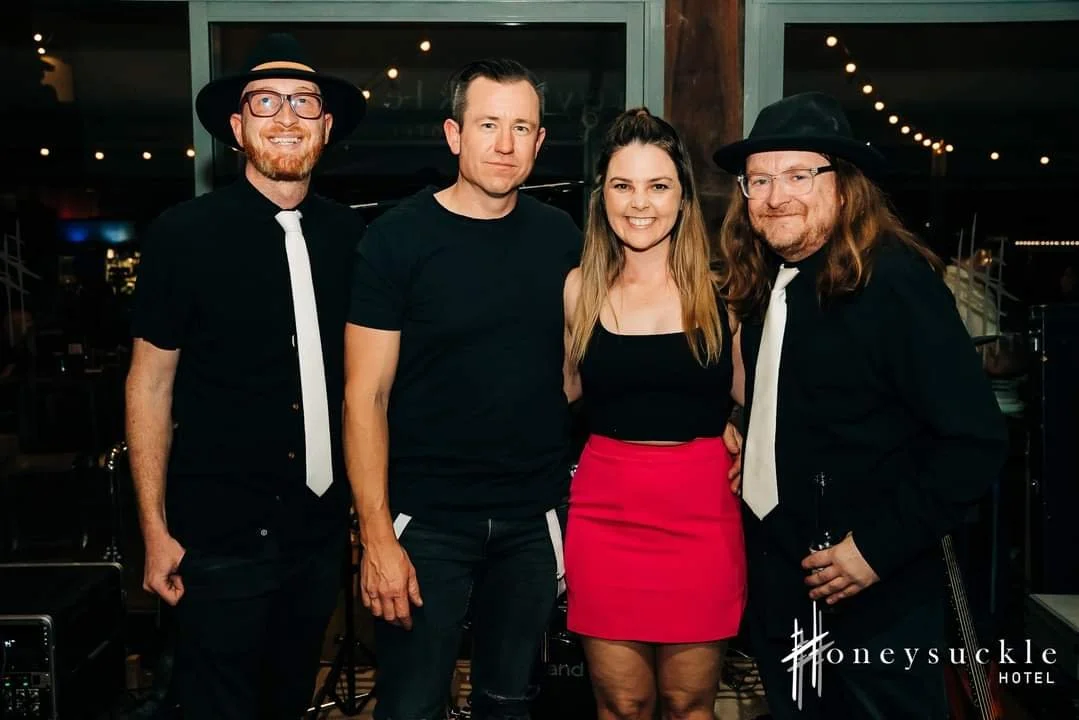 Four people posing together at an event inside HoneySuckle Hotel, with three men dressed in black and wearing hats, and a woman in a black top and red skirt. There are string lights in the background.