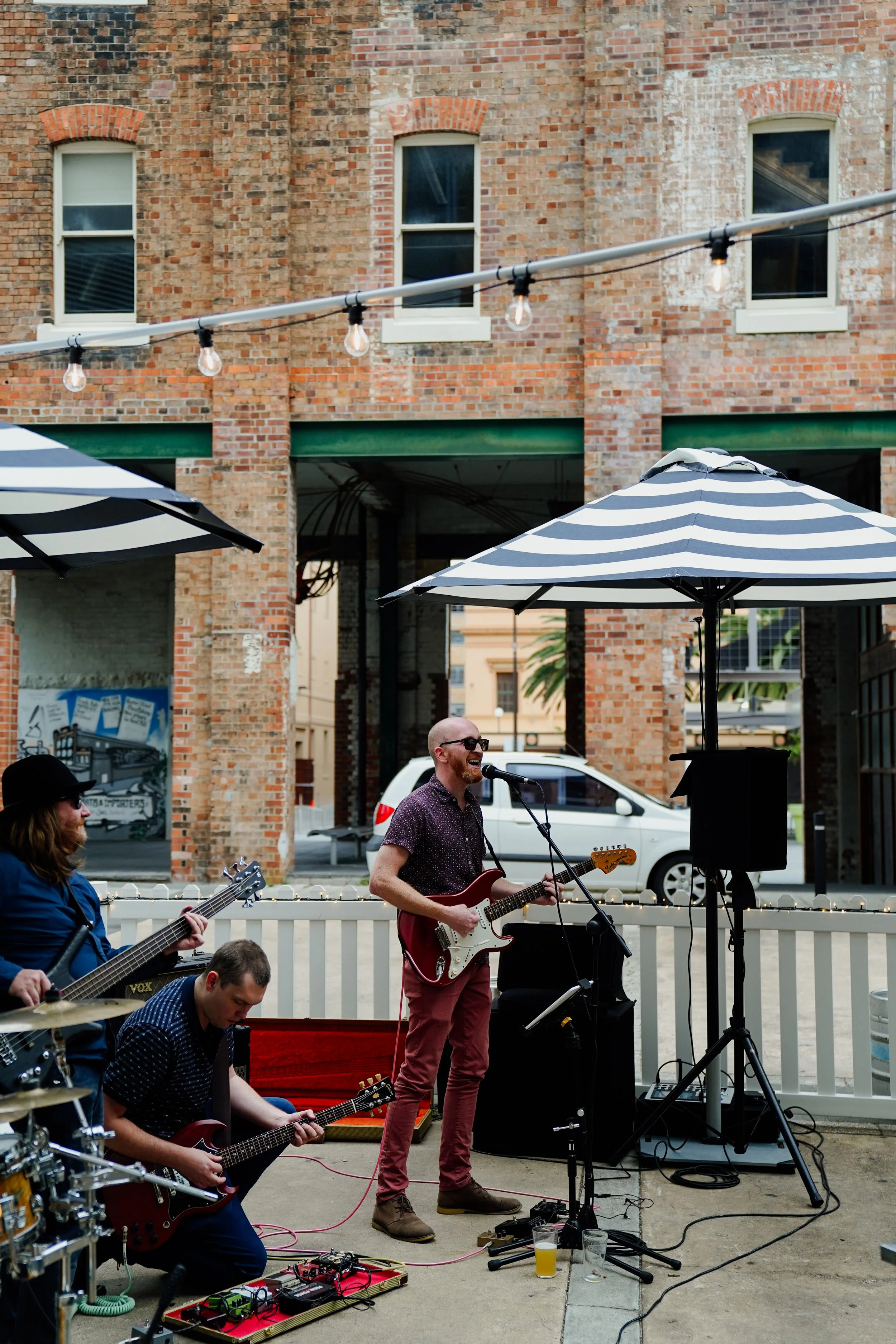 A musical band performs outdoors on a patio with brick walls, string lights, and striped umbrellas. One musician plays an electric guitar, singing into a microphone, while others play guitars and drums. There are drinks on the ground, and an audience