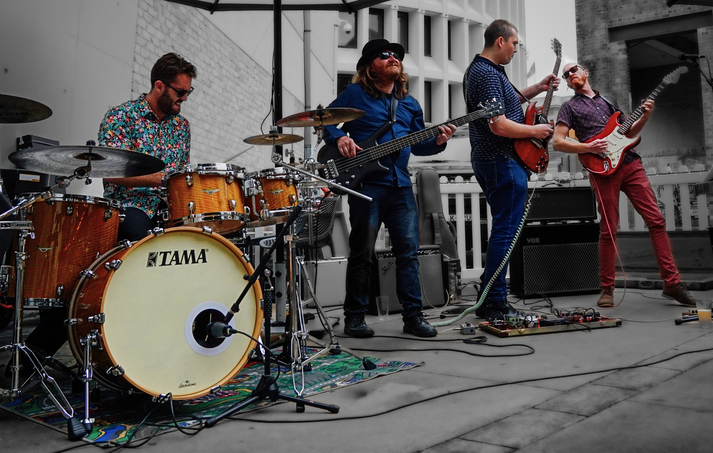 A four-member band performs on an outdoor stage, with one member playing drums, two on electric guitars, and one on bass guitar, standing in front of a brick and white building.
