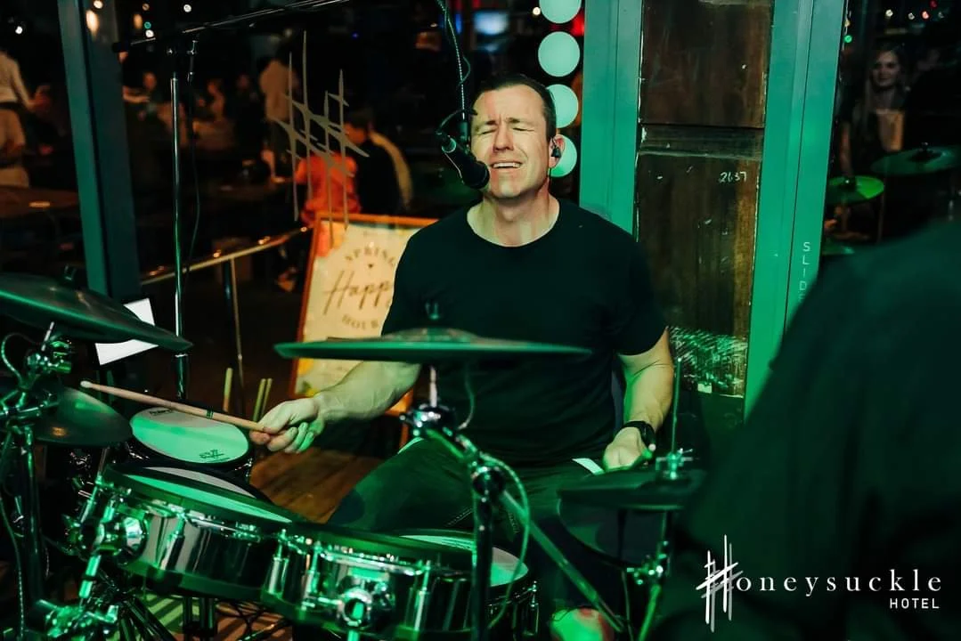 Man playing a drum set at a venue, singing with a passionate expression, with a microphone on his face, and a person in the foreground.