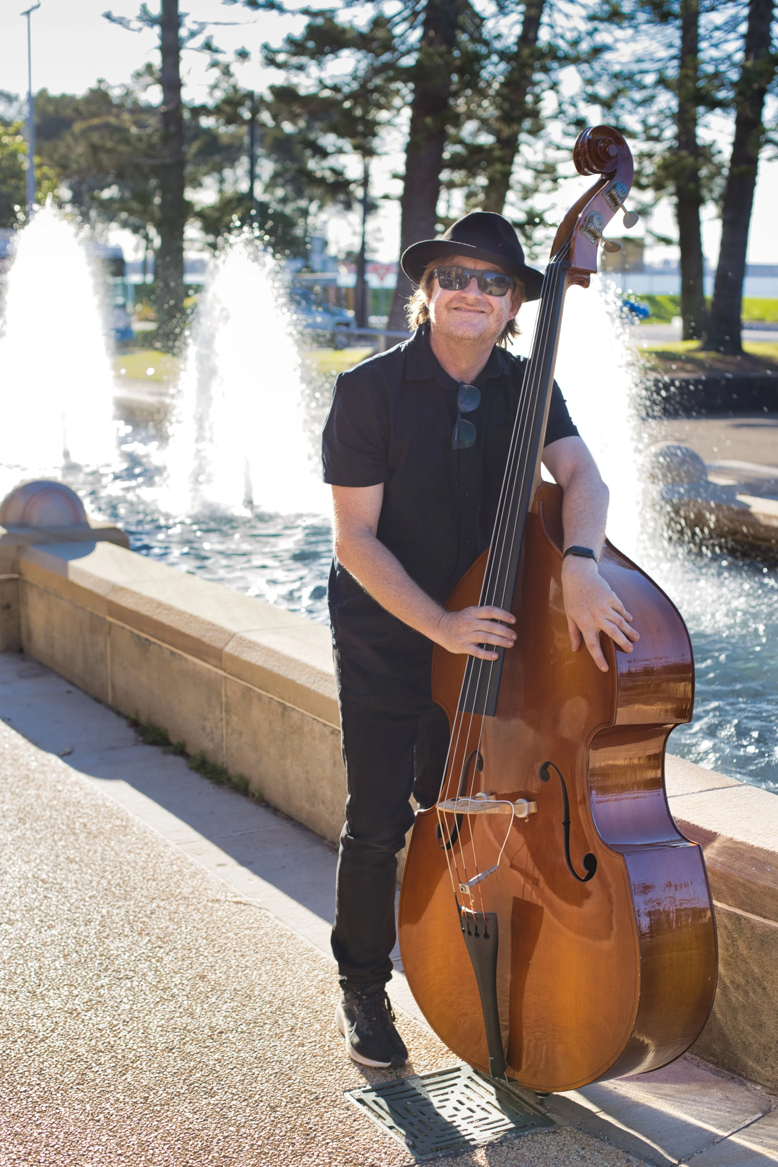 Man playing upright bass outdoors near a water fountain, wearing sunglasses, a wide-brimmed hat, and black clothing.