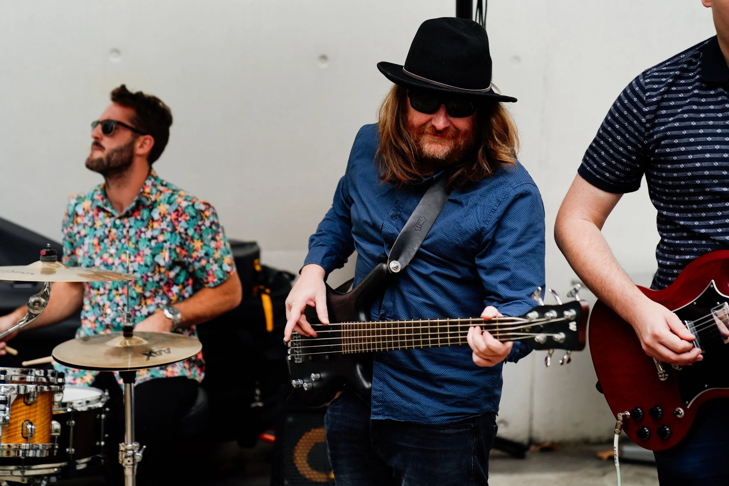Three musicians playing guitars and drums in a room. One man has long hair, a hat, sunglasses, and a beard. Another man has short hair, glasses, and a colorful shirt. The third person's arm and guitar are partially visible.
