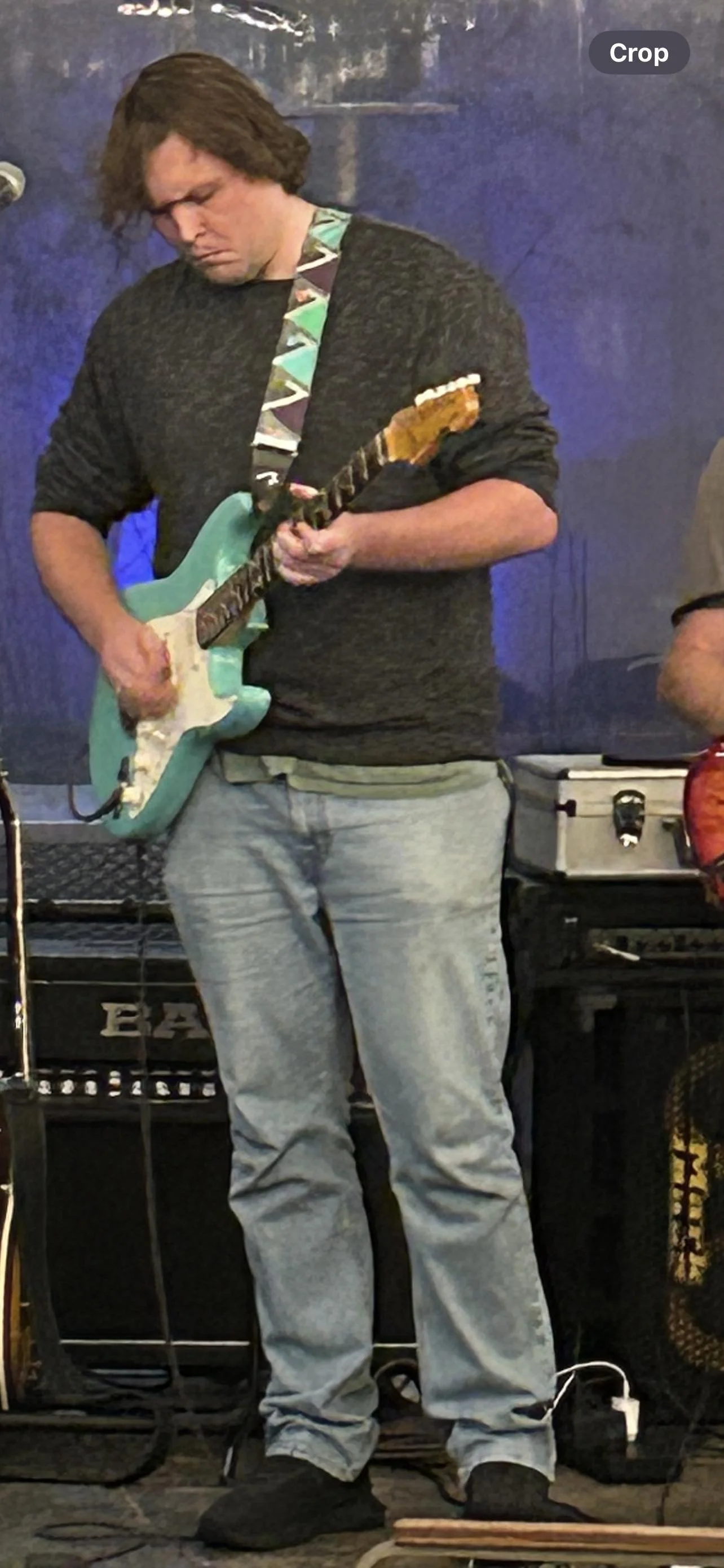 A person playing an electric guitar with a teal body and a multicolored strap, standing in front of amplifiers and equipment.