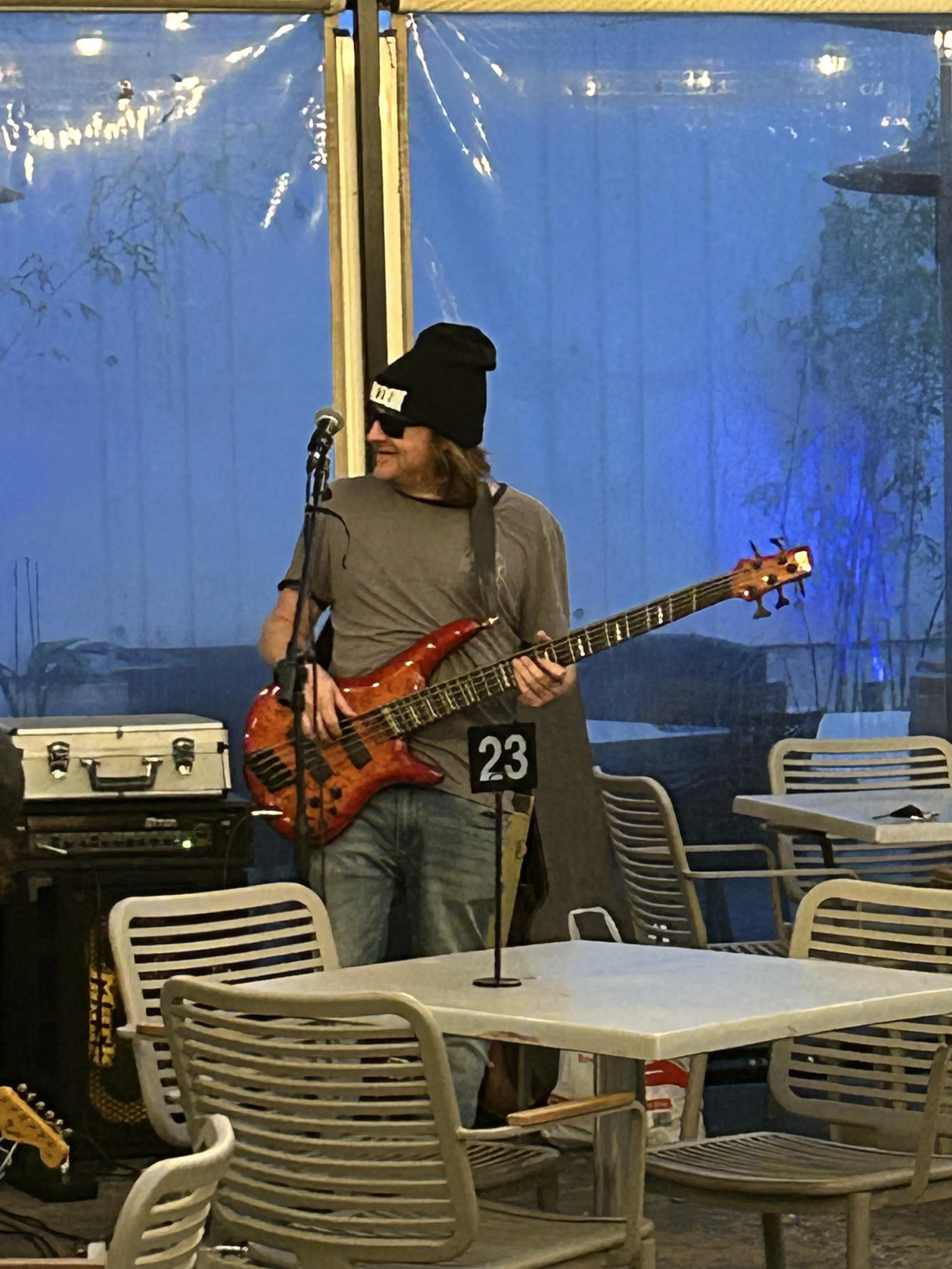 Musician playing an electric bass guitar with a guitar strap, wearing a black beanie and sunglasses, standing near a microphone, at an indoor venue with empty tables and chairs and a blue background window.