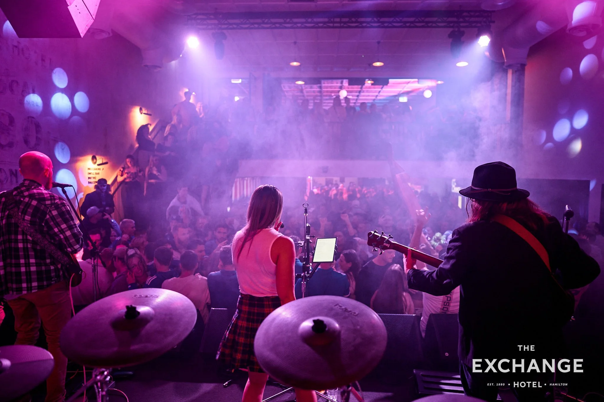 A live music performance at a nightclub with purple lighting and smoke effects. The band is on stage with a female singer, a guitarist, and other musicians. The audience is visible, with some people standing, enjoying the show. The venue is named The