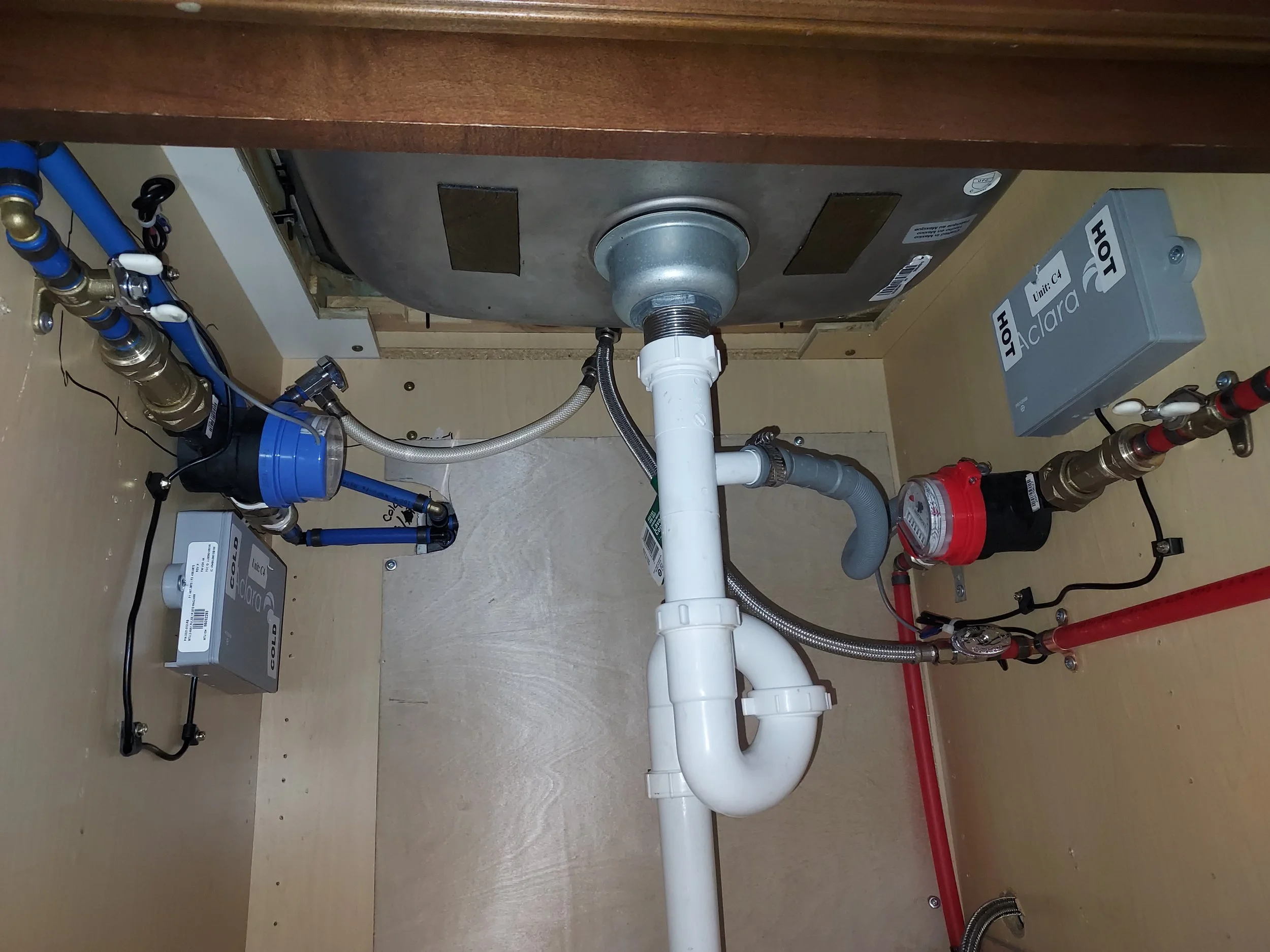 Under-sink plumbing with PVC pipes and water supply lines, electrical boxes labeled 'HOT' and 'COLD', and various valves and fittings.