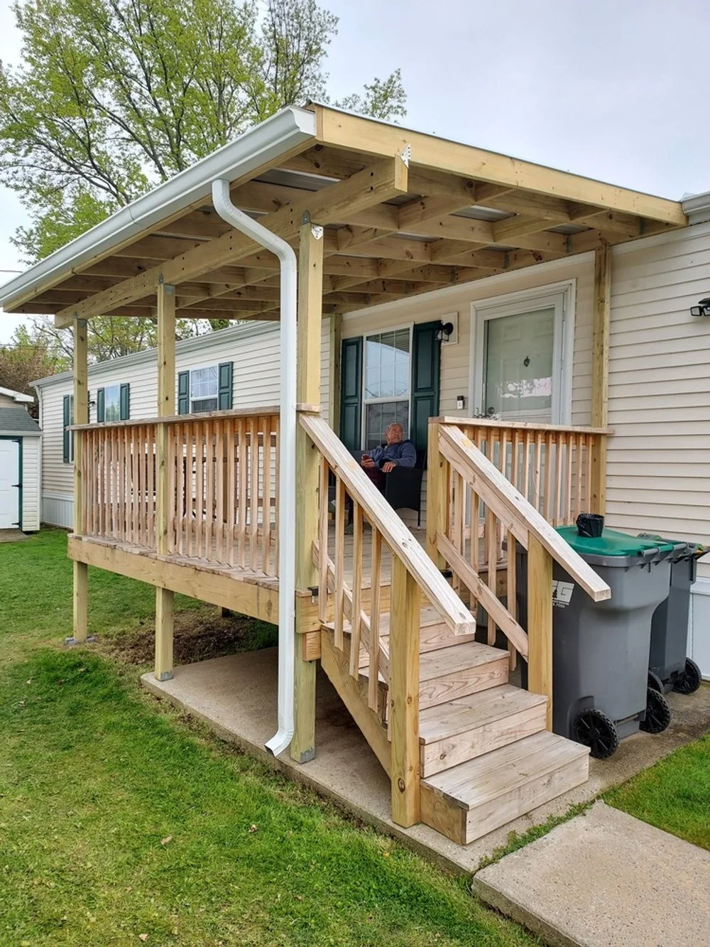 A newly built wooden porch with stairs and railing attached to a beige house. A man is sitting on a chair on the porch. Trash bins are nearby, and the porch is partially covered with an unfinished roof structure.