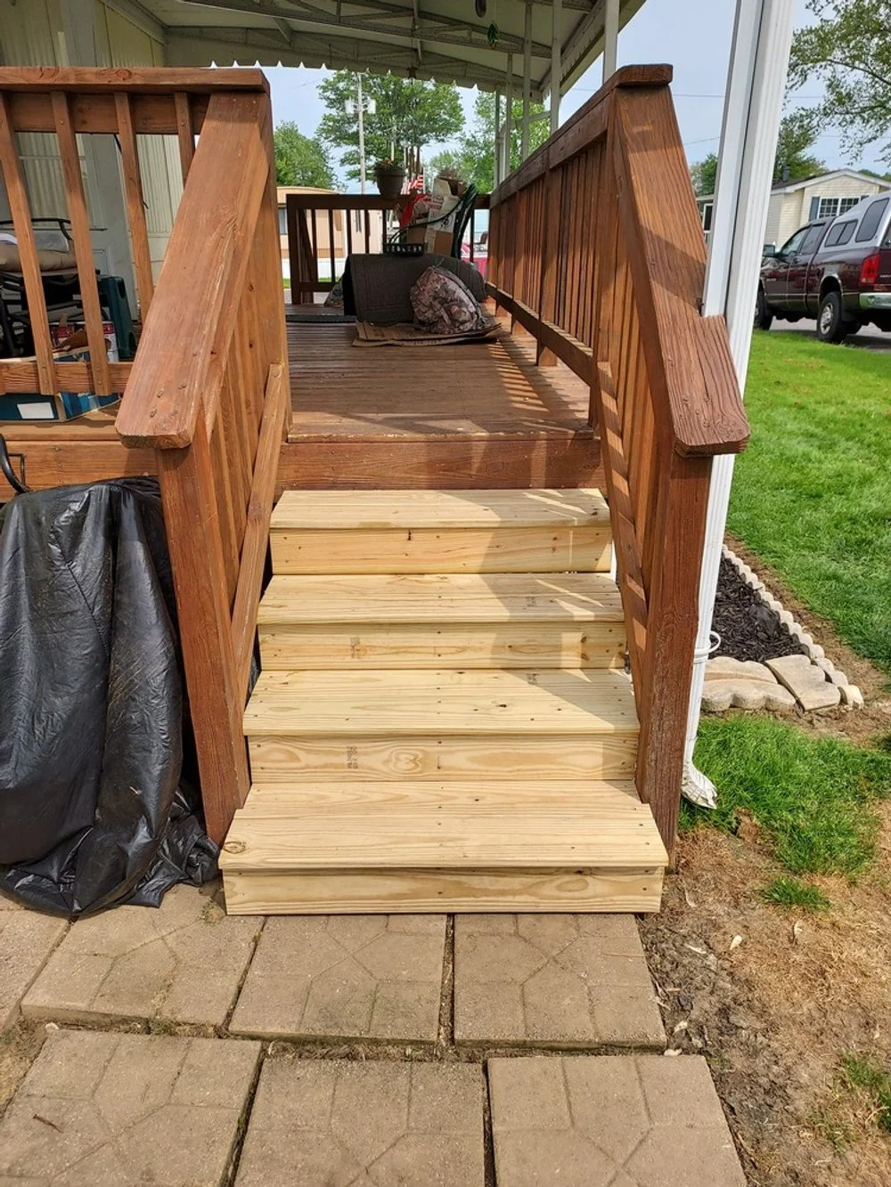 Wooden stairs leading up to a porch, with a two-tone railing and flooring, situated outdoors on a paved walkway with grass on the sides, in a residential area.