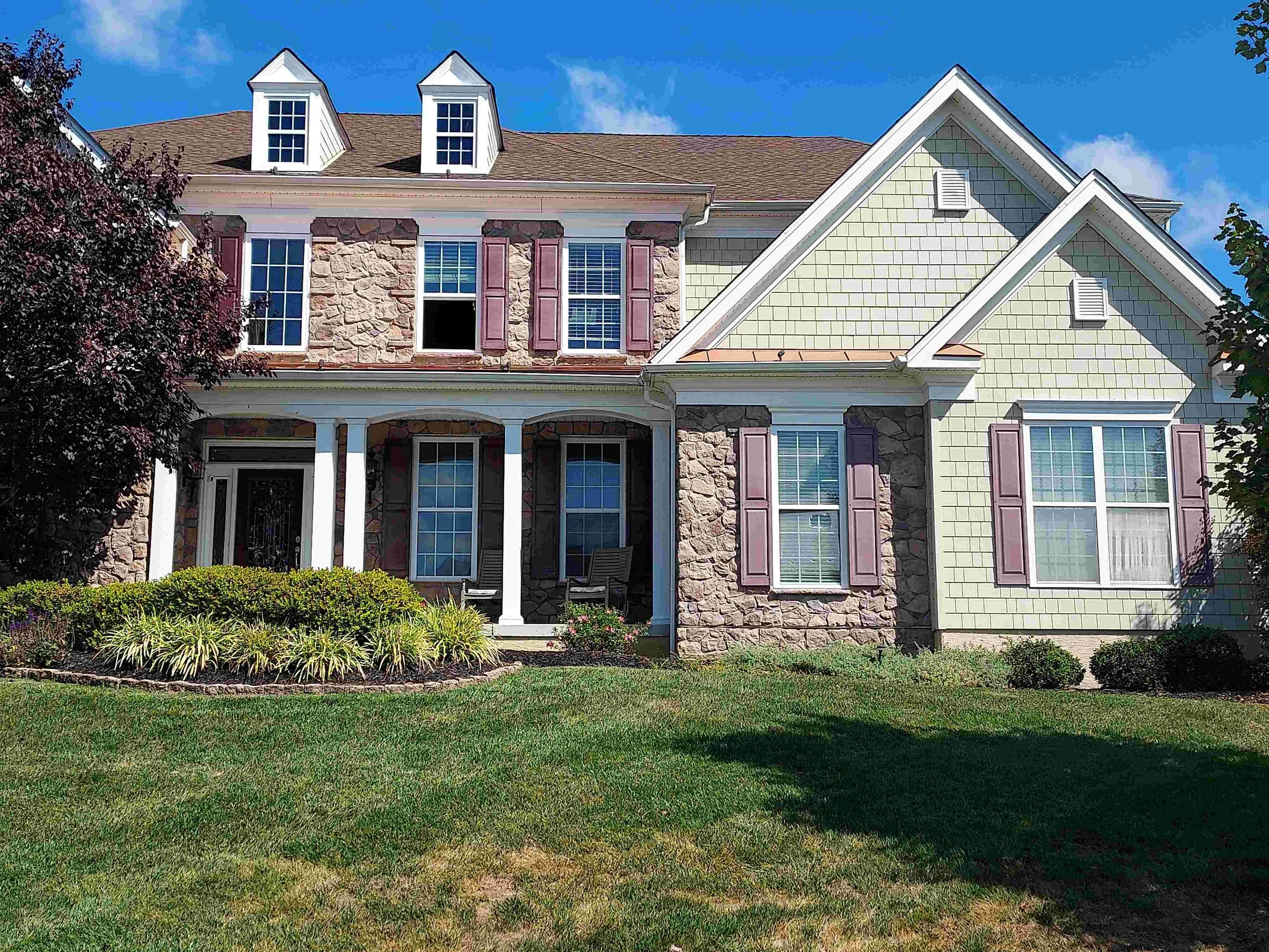 Front view of a two-story house with a stone facade, white siding, purple shutters, and a green shingled gable roof, under a partly cloudy blue sky.