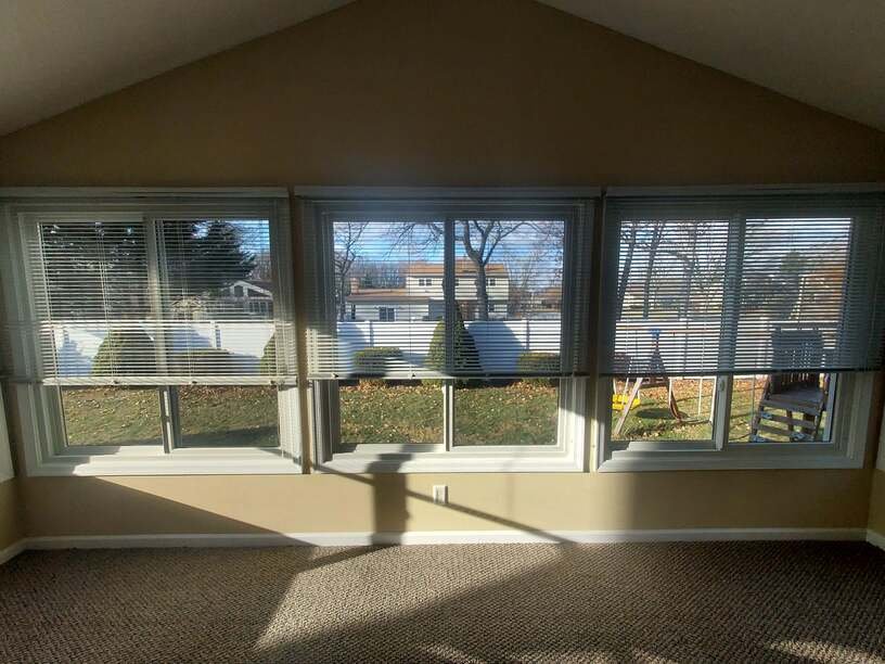 Interior view of a room with large windows and blinds, showing a yard and neighboring houses outside, with sunlight casting shadows.