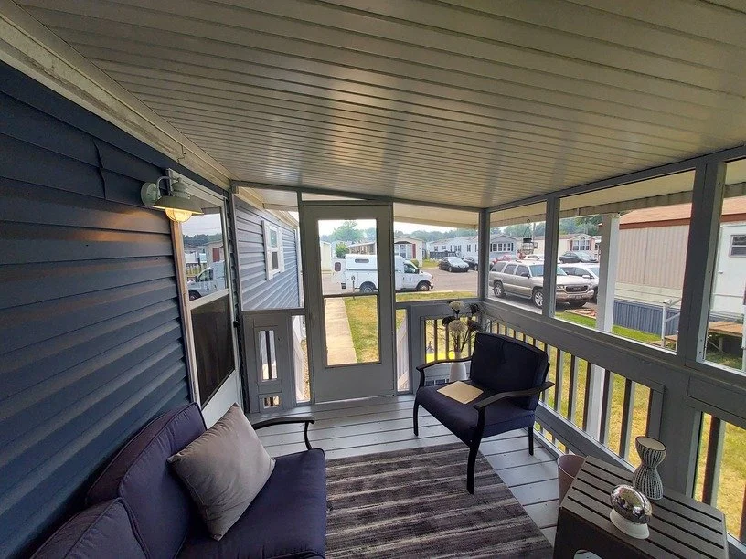 Enclosed porch with blue siding, seating area with a purple sofa and a black armchair, a small side table with a lamp and decorative items, and a view of mobile homes and parked cars outside.