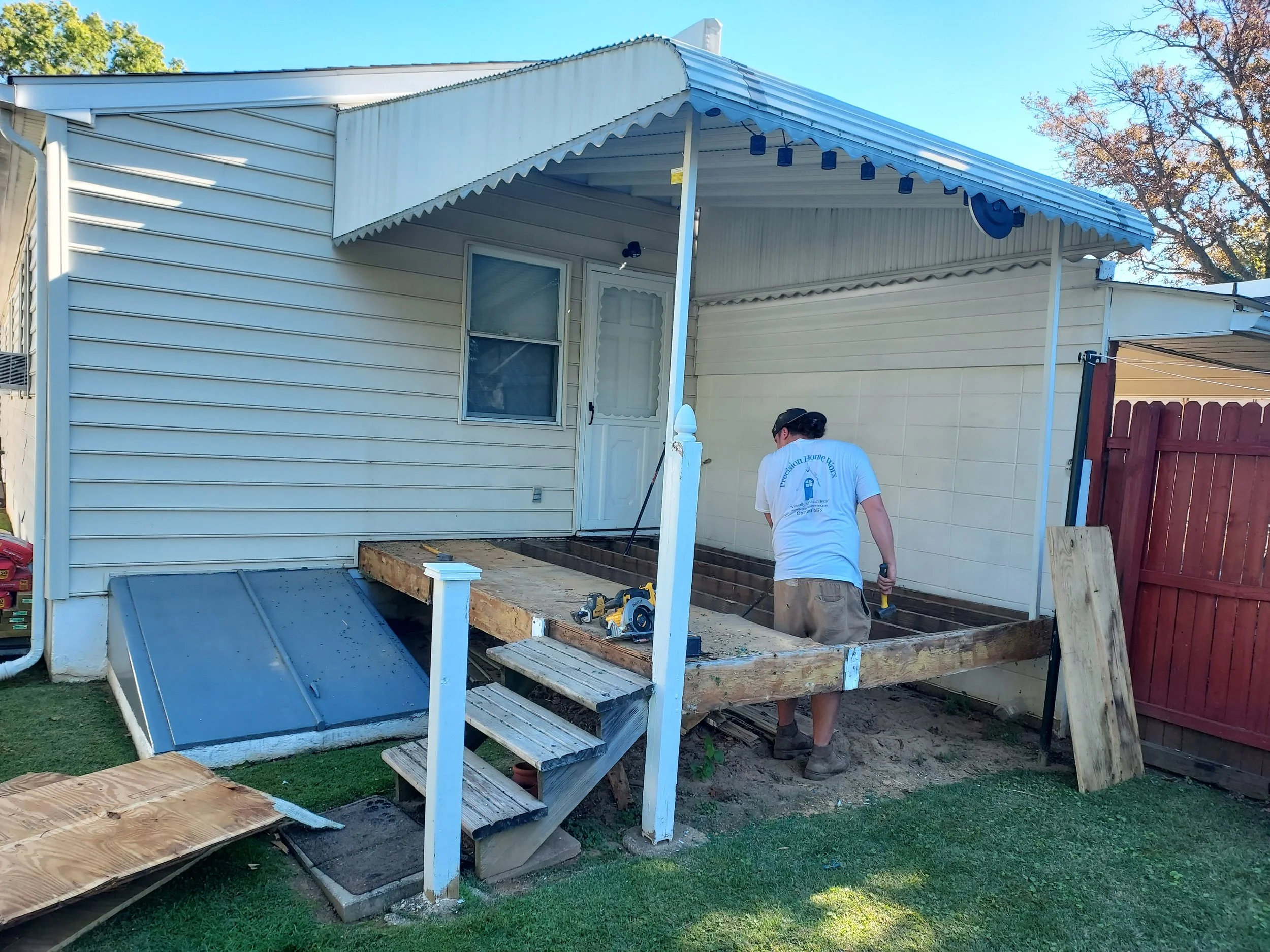 Person working on a construction project outside a house, installing or repairing the porch, with exposed wooden framework, tools around, and a red fence nearby.