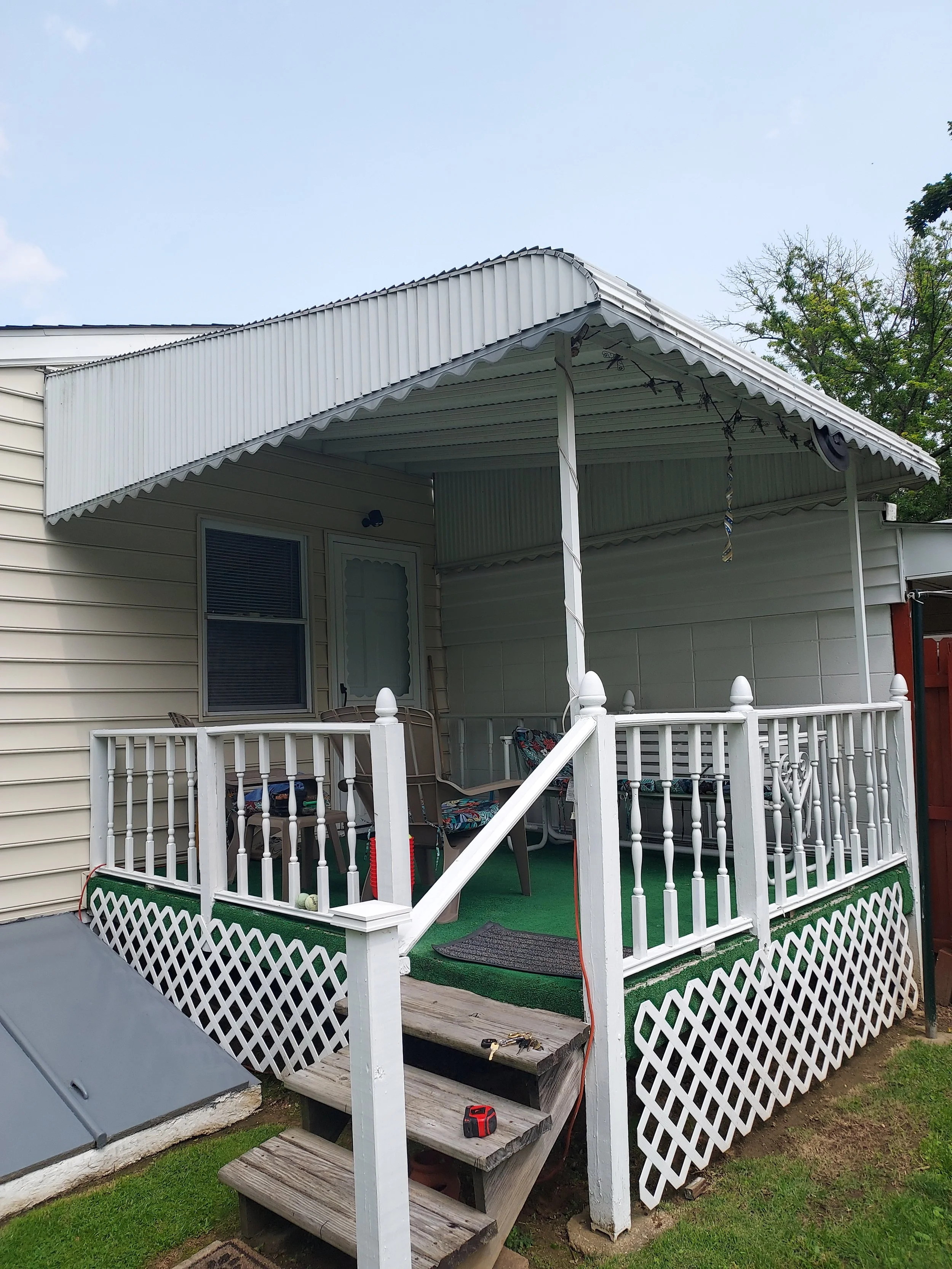 A house with a porch, white railing and lattice, and a roofed section with hanging string lights; construction tools are on the steps.