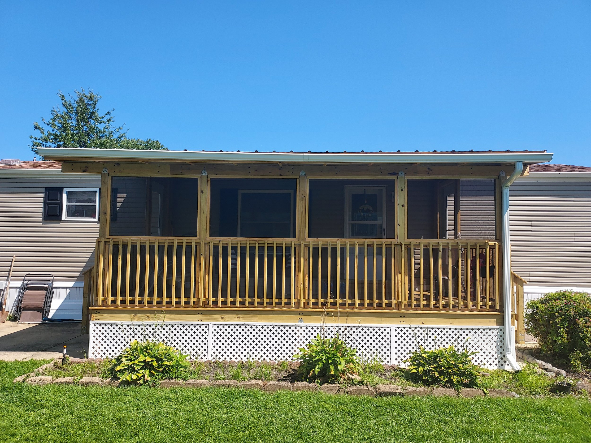 Backyard view of a house with a newly built screened porch with a wooden railing, beige siding, and a shingled roof, green plants in the foreground, and a tree and blue sky in the background.
