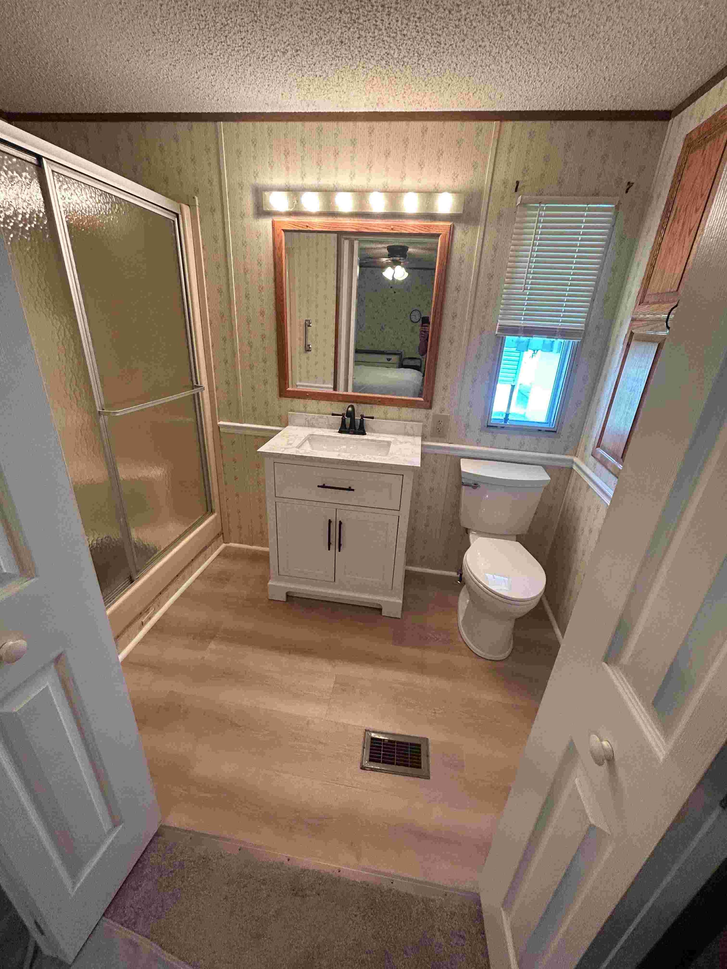 Small bathroom with shower stall, white vanity with sink, mirror, toilet, small window with blinds, wood flooring, and heating vent.