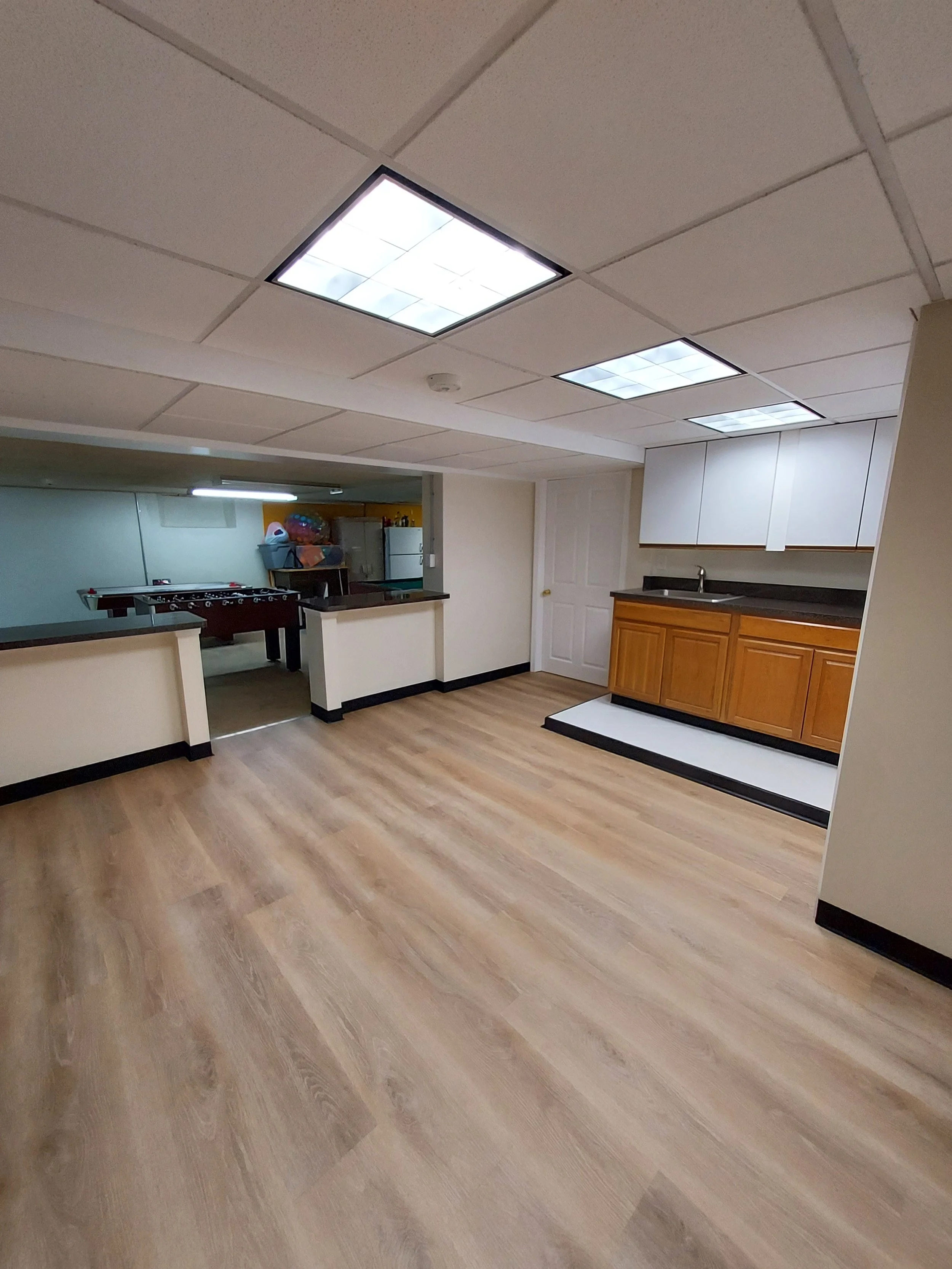 A basement room with beige drop ceiling, fluorescent lights, wooden flooring, small kitchenette area with white cabinets and a sink, door, and a foosball table against the wall.