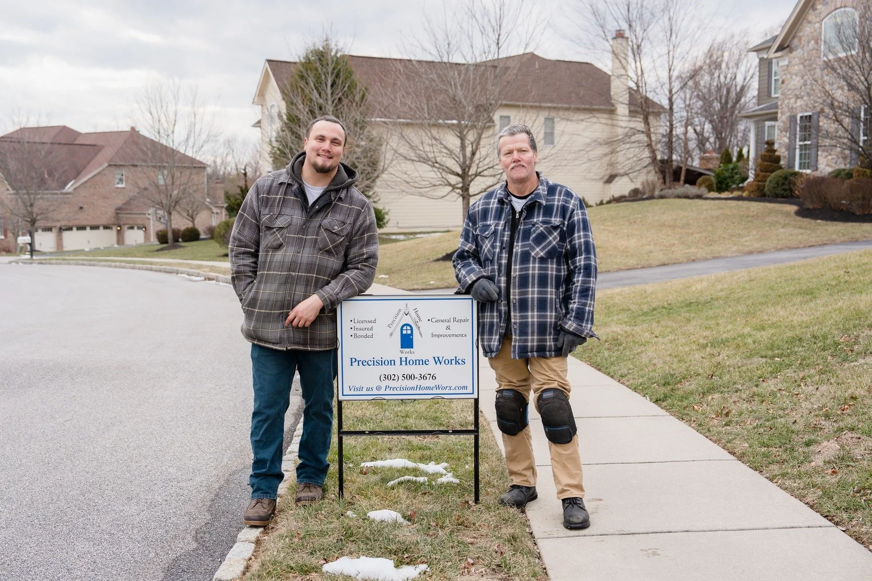Two men standing outside a house, one wearing a plaid jacket and the other in a flannel shirt, next to a sign for Precision Home Works, a home repair and improvement service.