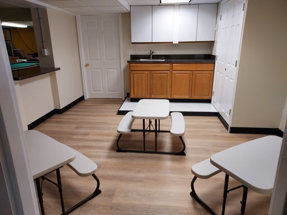 A small kitchen area with wooden cabinets, a black countertop, and a sink. There are three white folding tables with attached benches on a light wood floor. A white door is visible in the background.