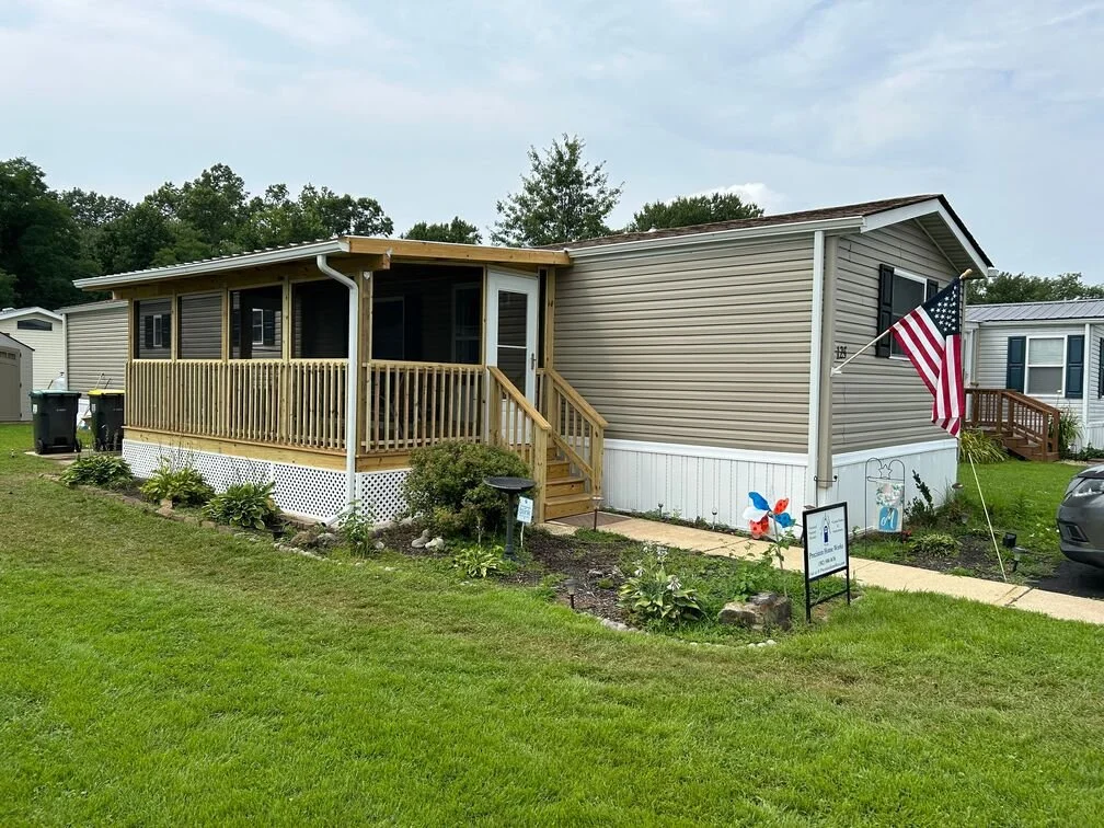 A manufactured home with a new wooden porch and stairs, surrounded by a lawn with plants and garden decor, an American flag on a pole, and a sign in front.