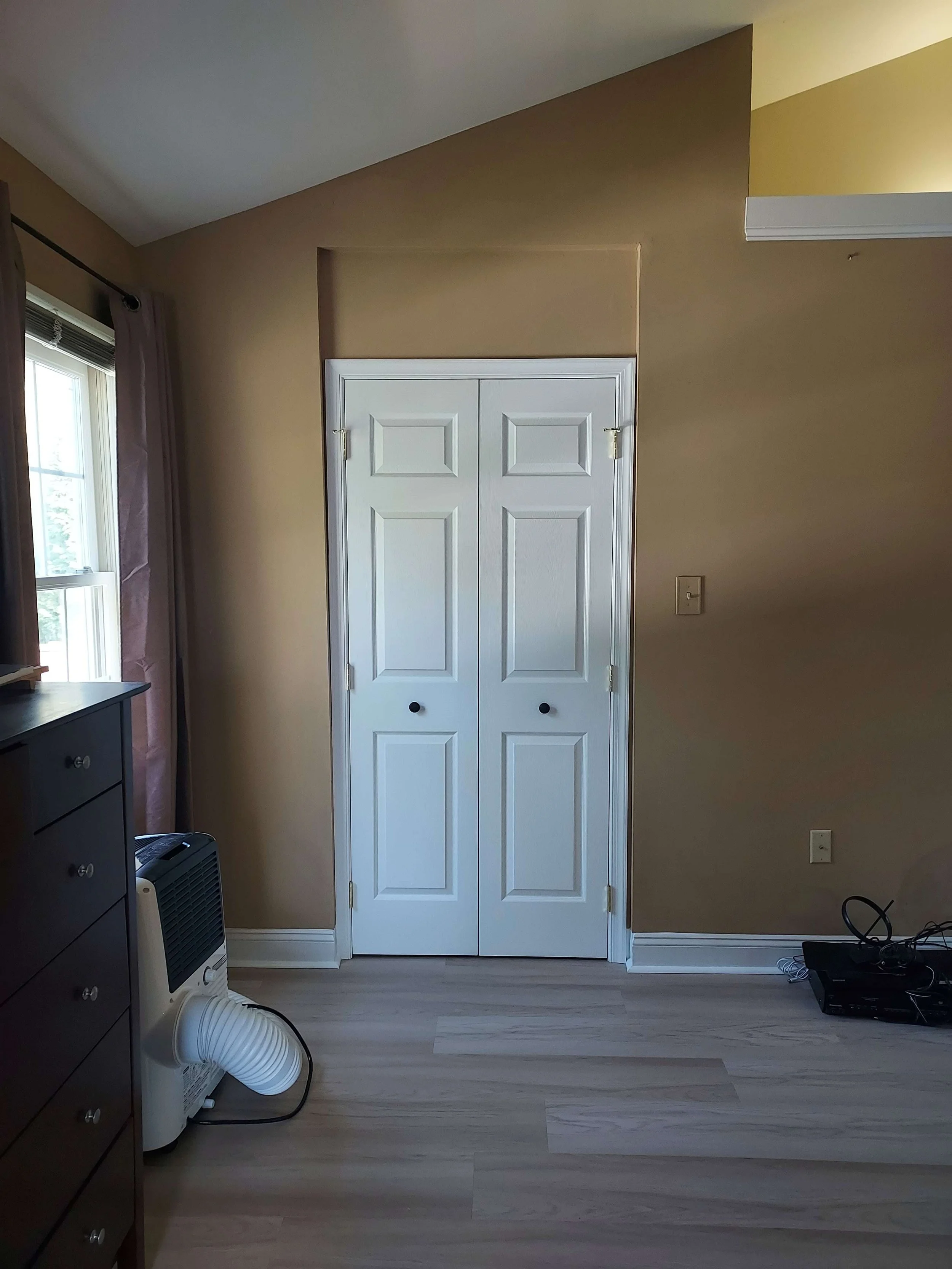 A room with a double closet door, beige walls, a window with curtains, a heater, and various electronic devices on the floor.