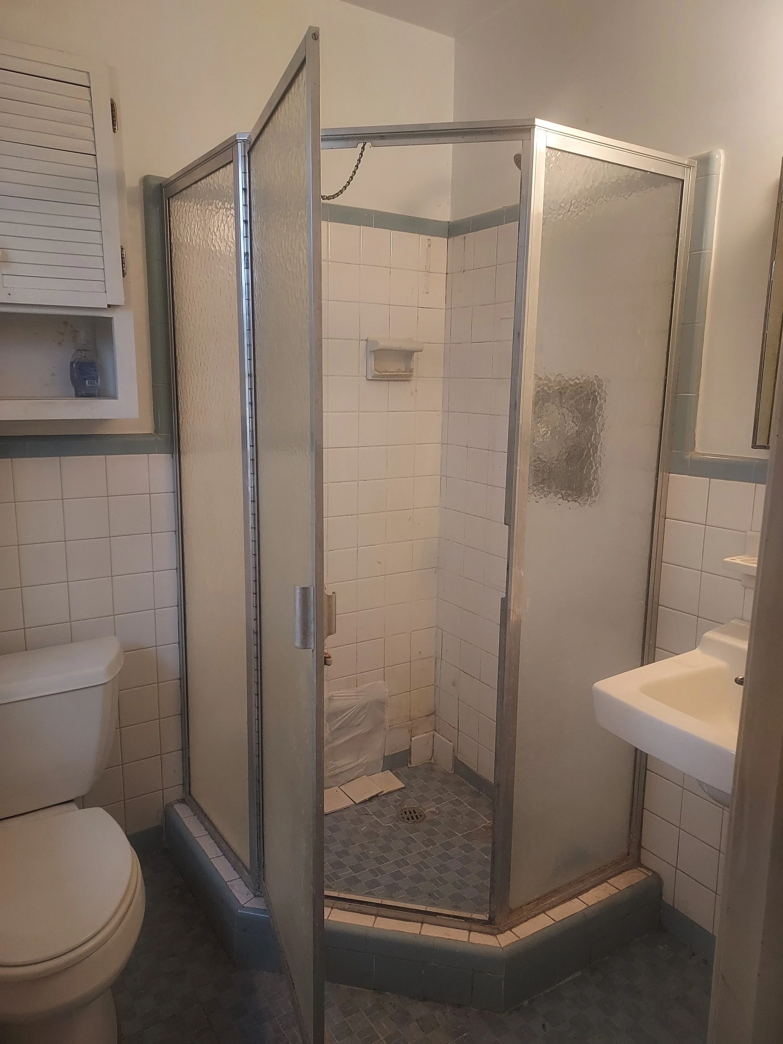 A small bathroom with a shower stall, toilet, and sink. The shower has frosted glass doors and white tile walls. The toilet and sink are white, and the floor has small gray tiles.