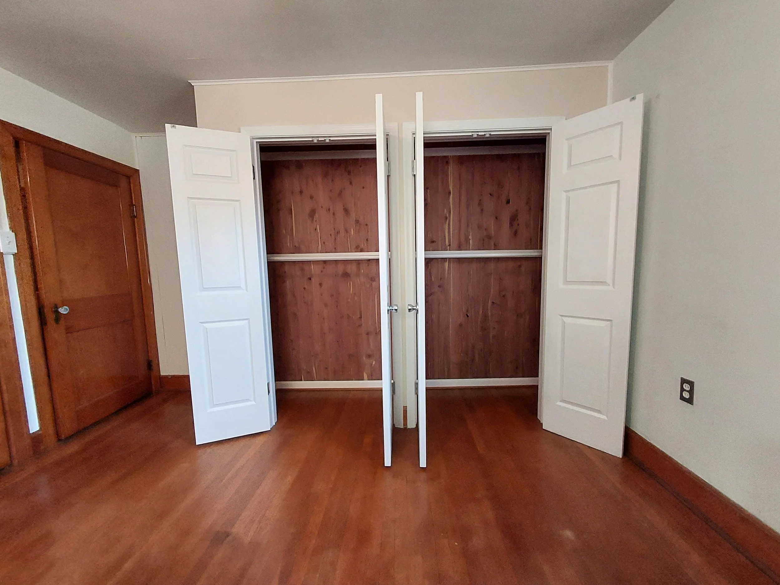 An empty closet with white folding doors, showing a wooden interior, in a room with hardwood floors and light-colored walls.