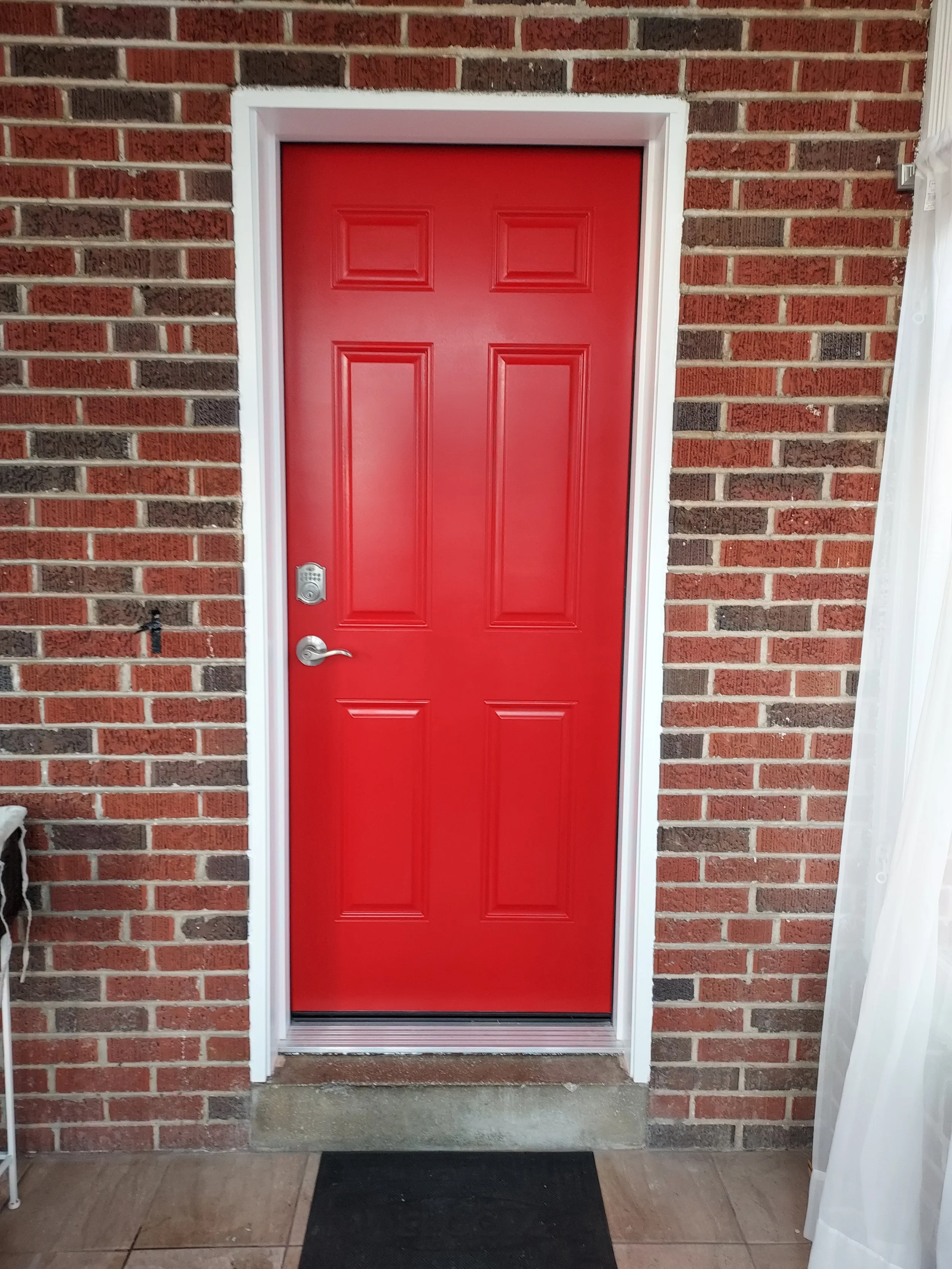 Red door with white trim housing a keypad lock, set in a brick wall with beige and dark brown bricks. Dark doormat in front, beige tiled floor, and partial white curtain on the right.