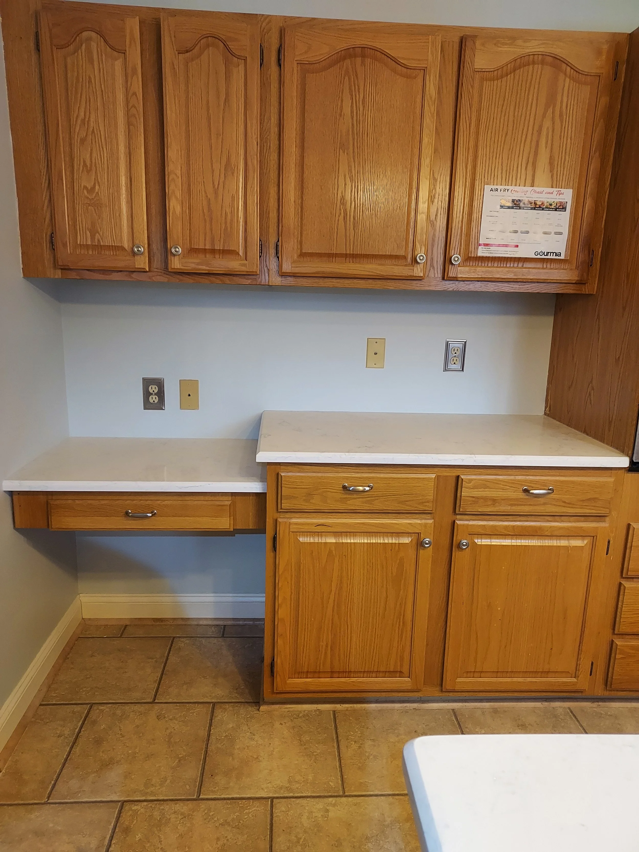 Kitchen cabinets above a countertop with a small chart attached, electrical outlets on the wall, and tan tile flooring.