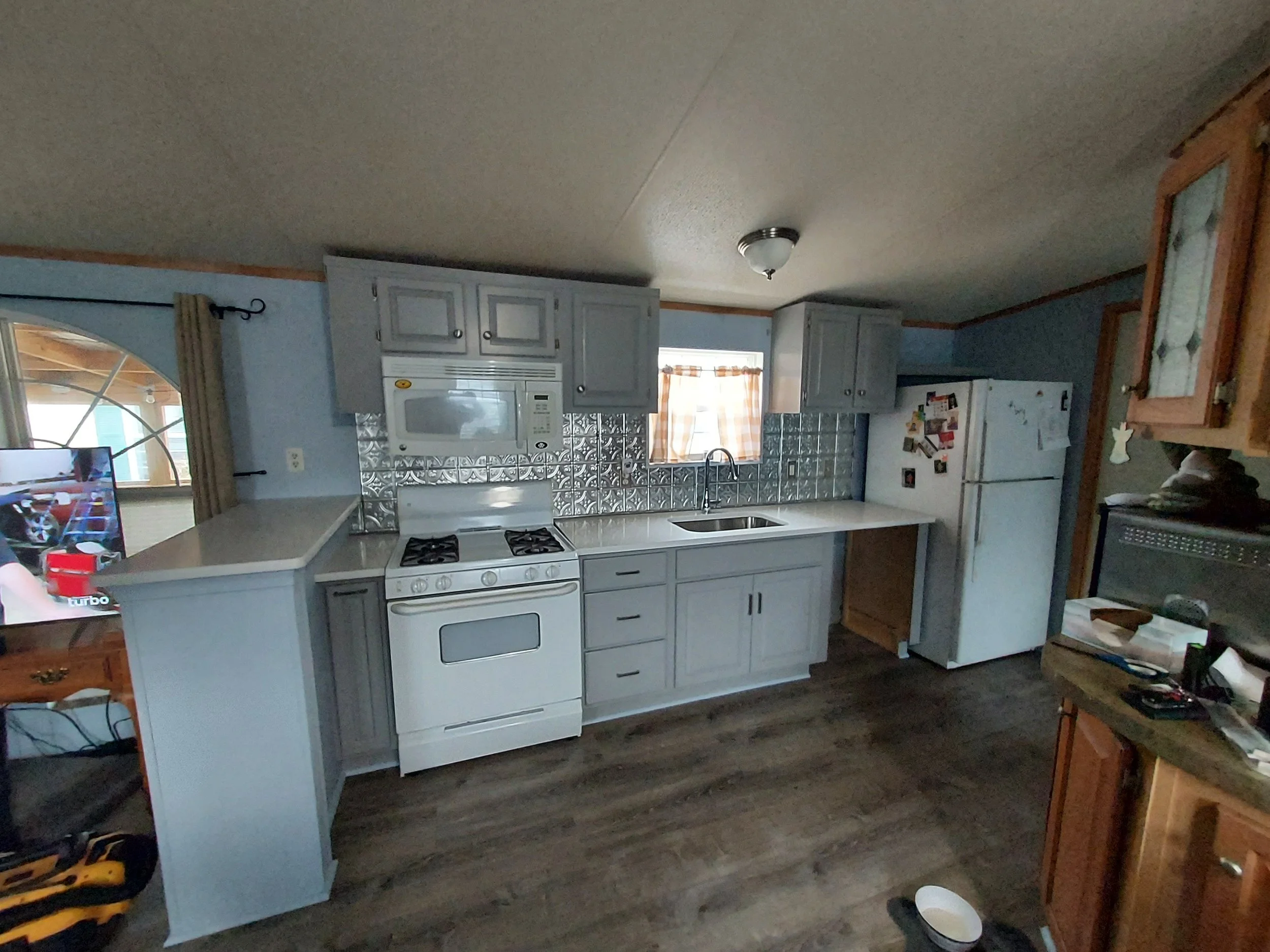 Kitchen with light gray cabinets, white appliances, and a stainless steel sink under a window with checkered curtains. There is a stove and microwave, and various items on countertops and a nearby table. The flooring is dark wood.