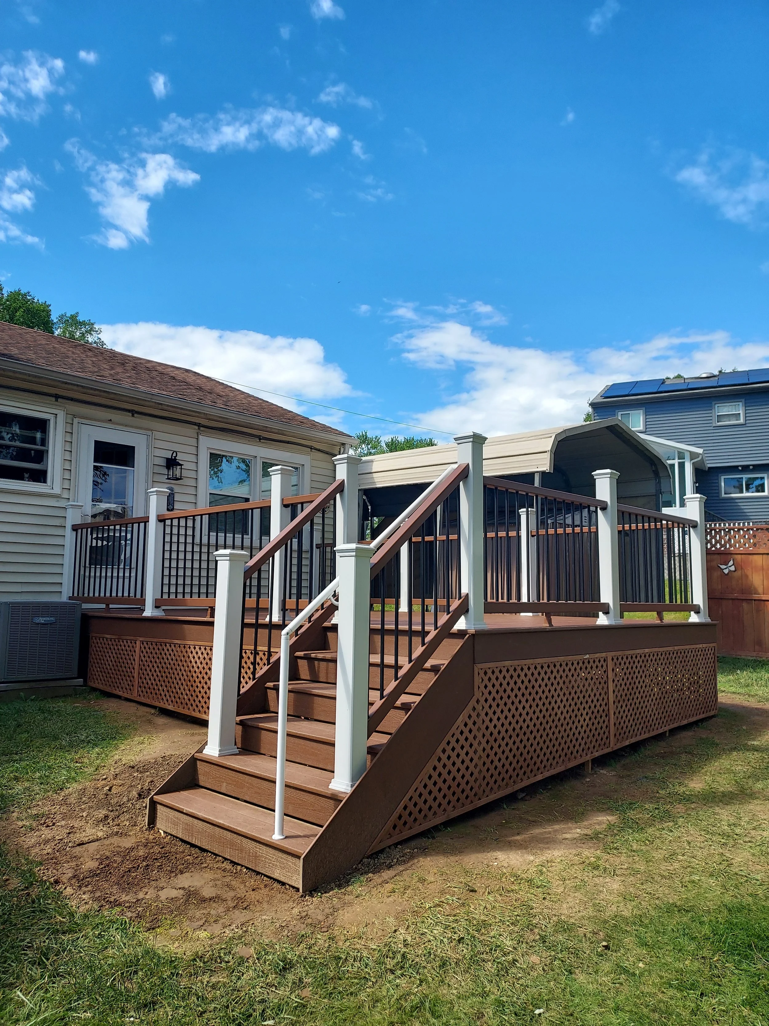 A backyard wooden deck with stairs, white railings, and lattice skirting, attached to a beige house with brown shingle roof. There is a fence on the right side, and the yard has grass and patches of dirt. A neighboring blue house with solar panels is visible in the background under a partly cloudy sky.
