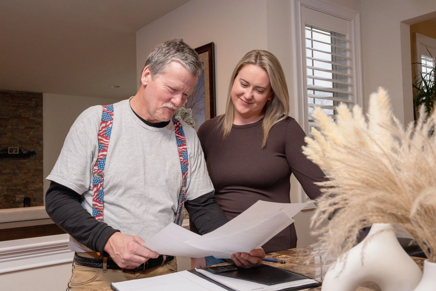 A man and a woman looking at papers together in a kitchen with a vase of dried flowers on the counter.