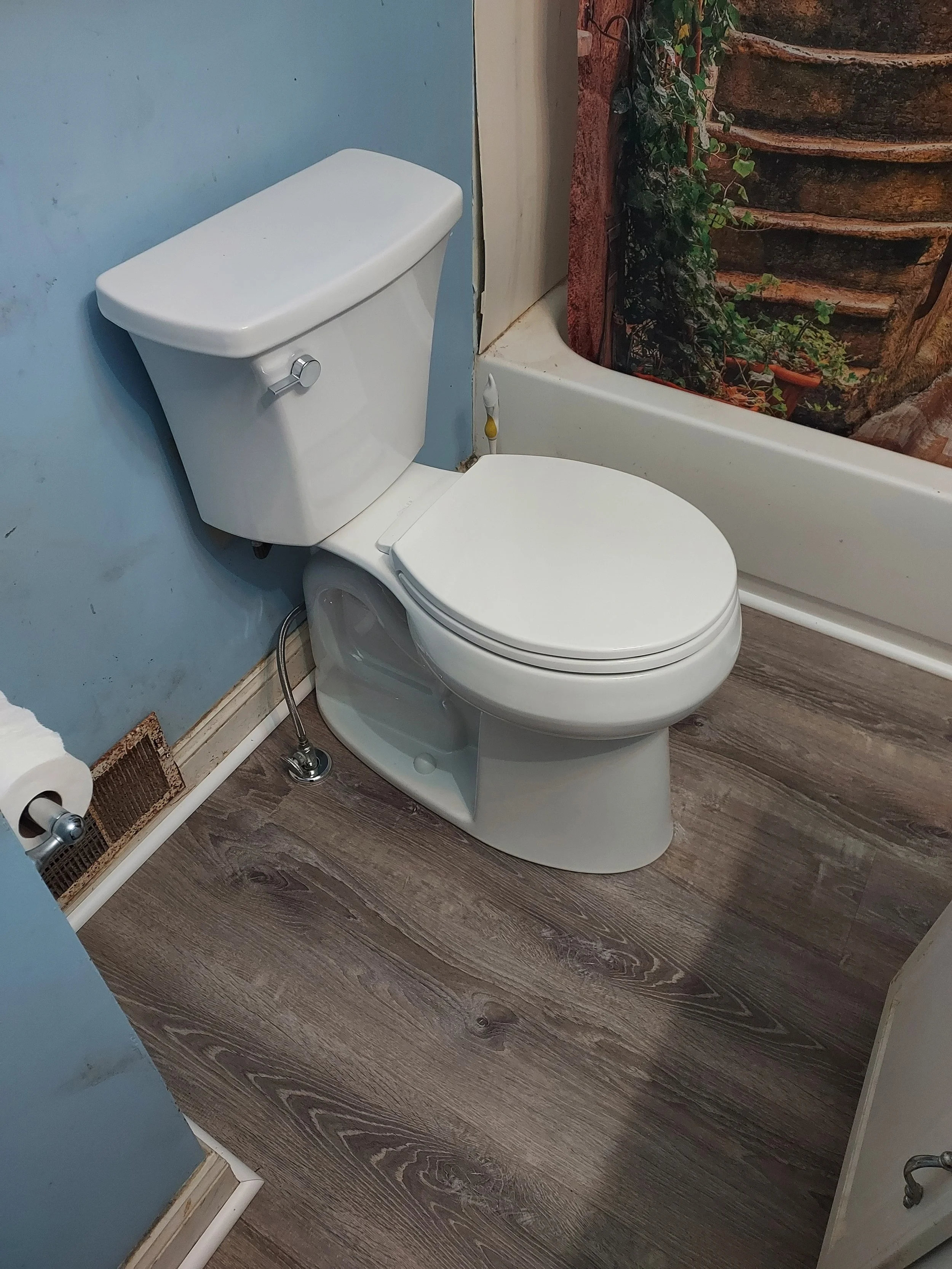 A bathroom with a white toilet and flush handle, blue wall, partial view of wooden floor, and a shower curtain with a nature scene of trees and plants.