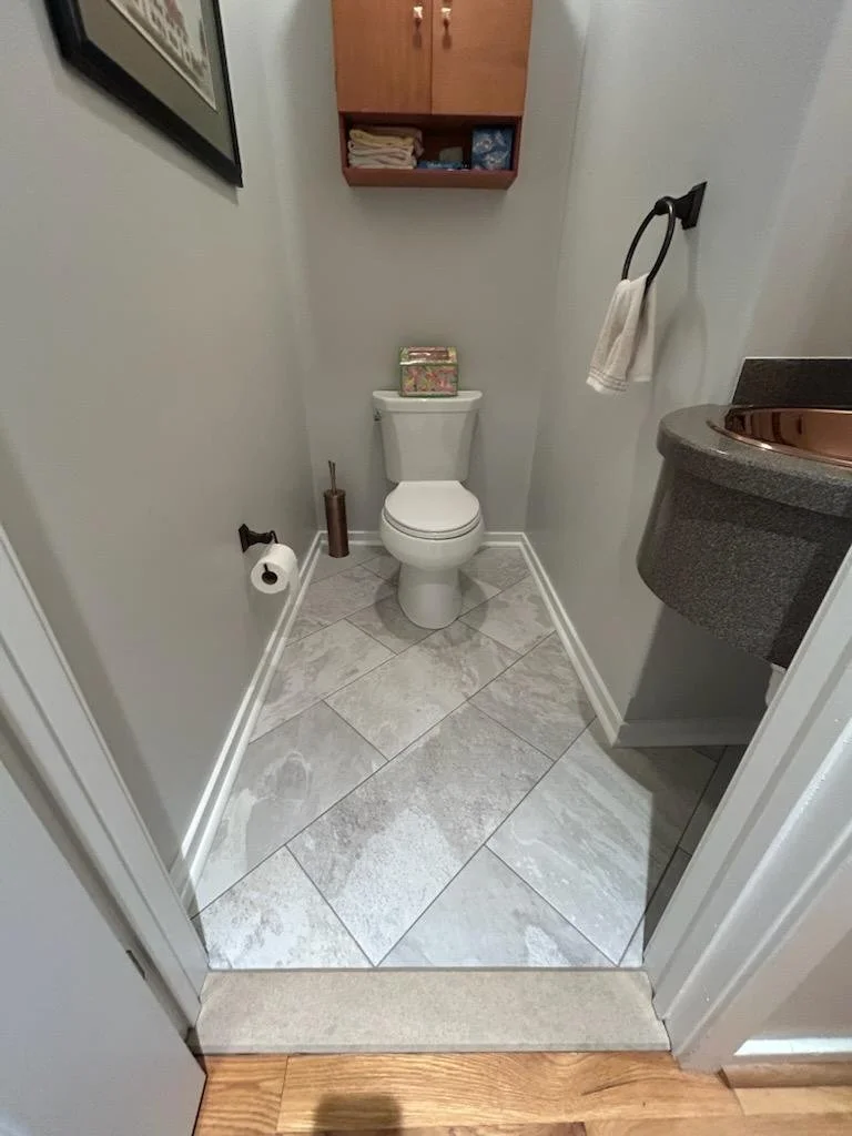 Small bathroom with a white toilet, wooden wall cabinet above with towels and supplies, toilet paper on holder on left wall, brown toilet brush holder next to the toilet, small sink with a copper-colored basin on the right, towel ring with a towel on the right wall, framed picture on the left wall, tile floor, doorframe at the bottom.