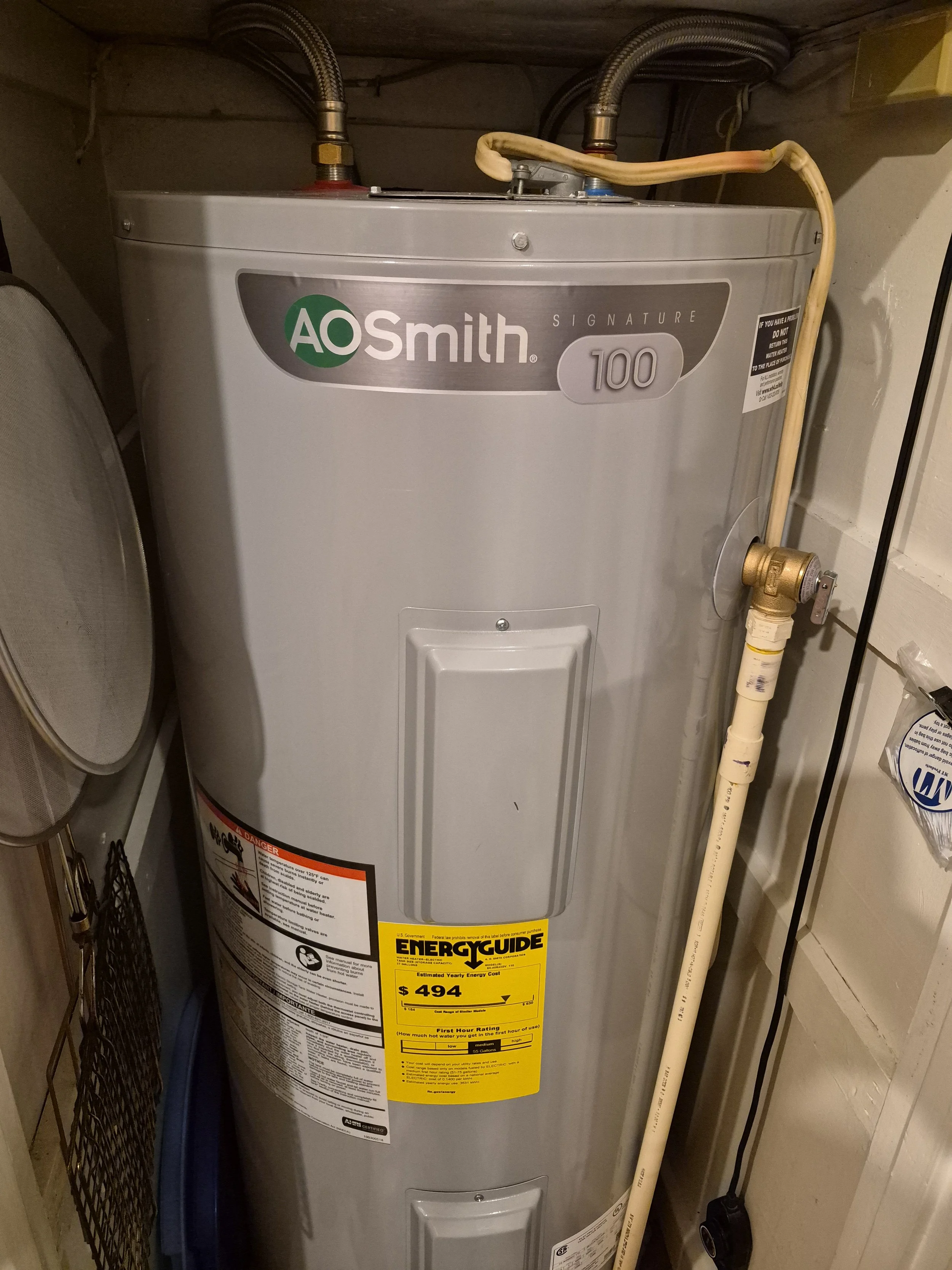 A metallic A.O. Smith Signature 100 water heater in a utility closet with pipes, hoses, and an energy guide label.