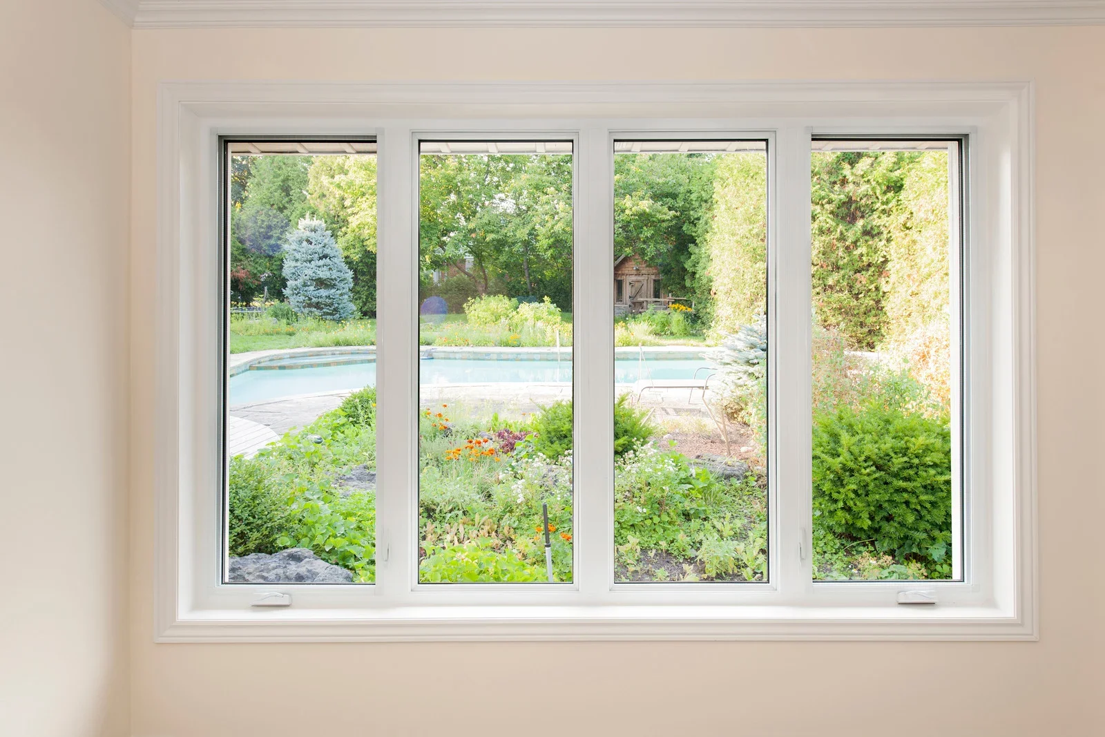 View through a large white-framed window showing a backyard with a swimming pool, green trees, bushes, and a garden with colorful flowers.