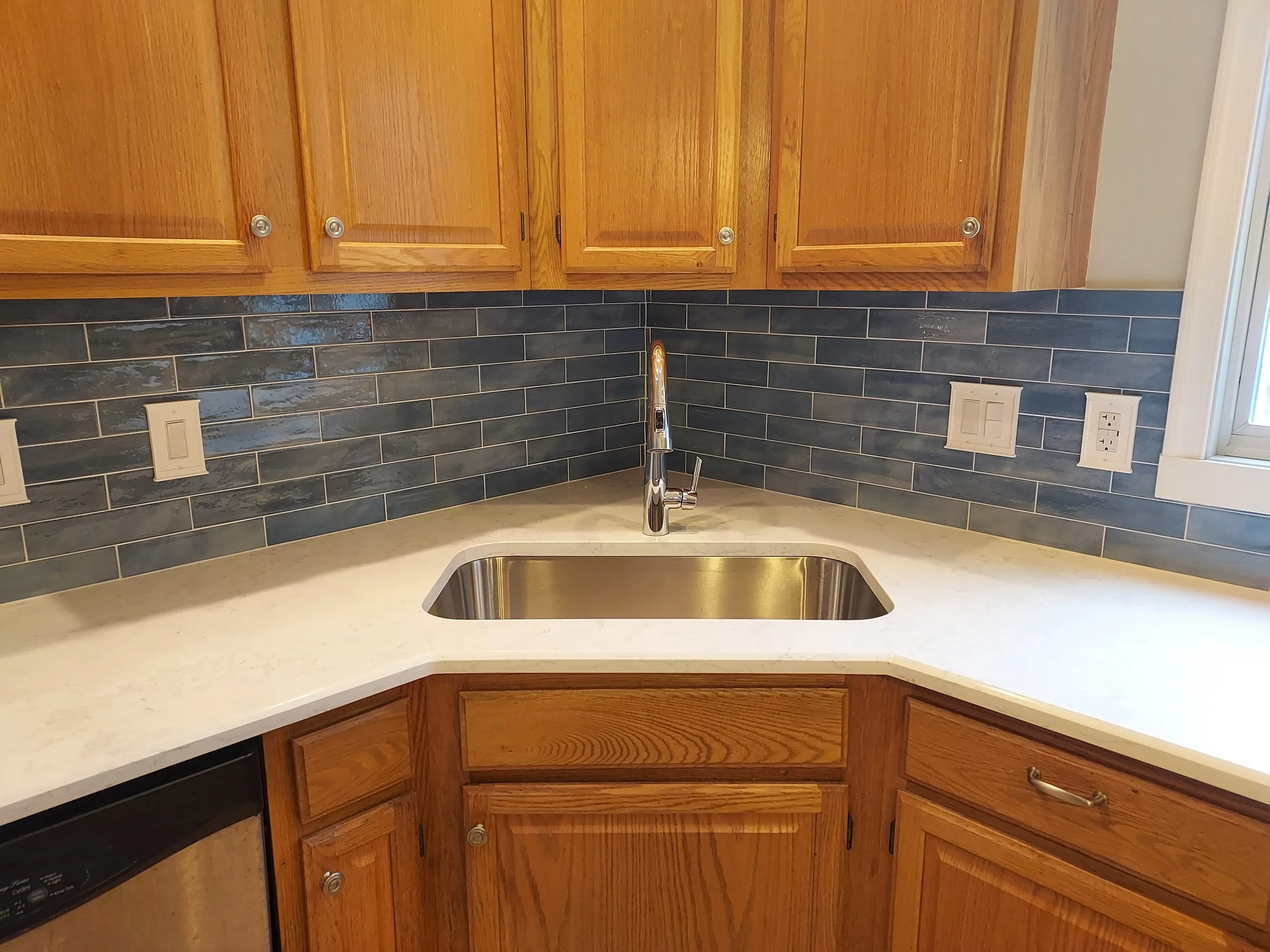 Kitchen with wooden cabinets, black tile backsplash, and a white countertop with a stainless steel sink and faucet.
