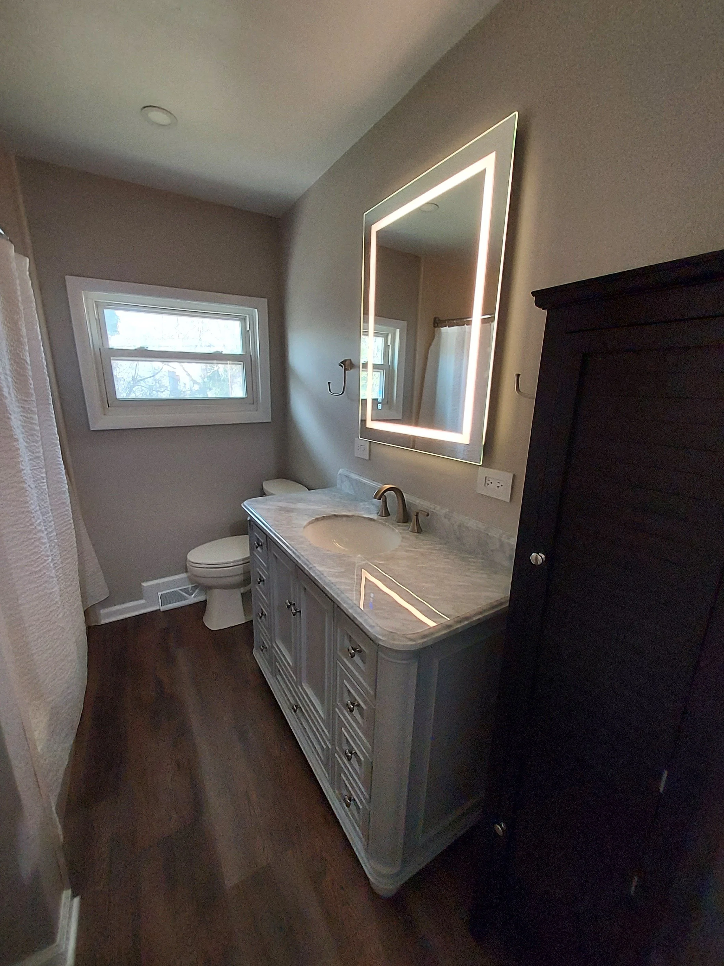 Bathroom with a window, a white vanity with a marble countertop, a mirror with LED lights, a dark cabinet, and a toilet.
