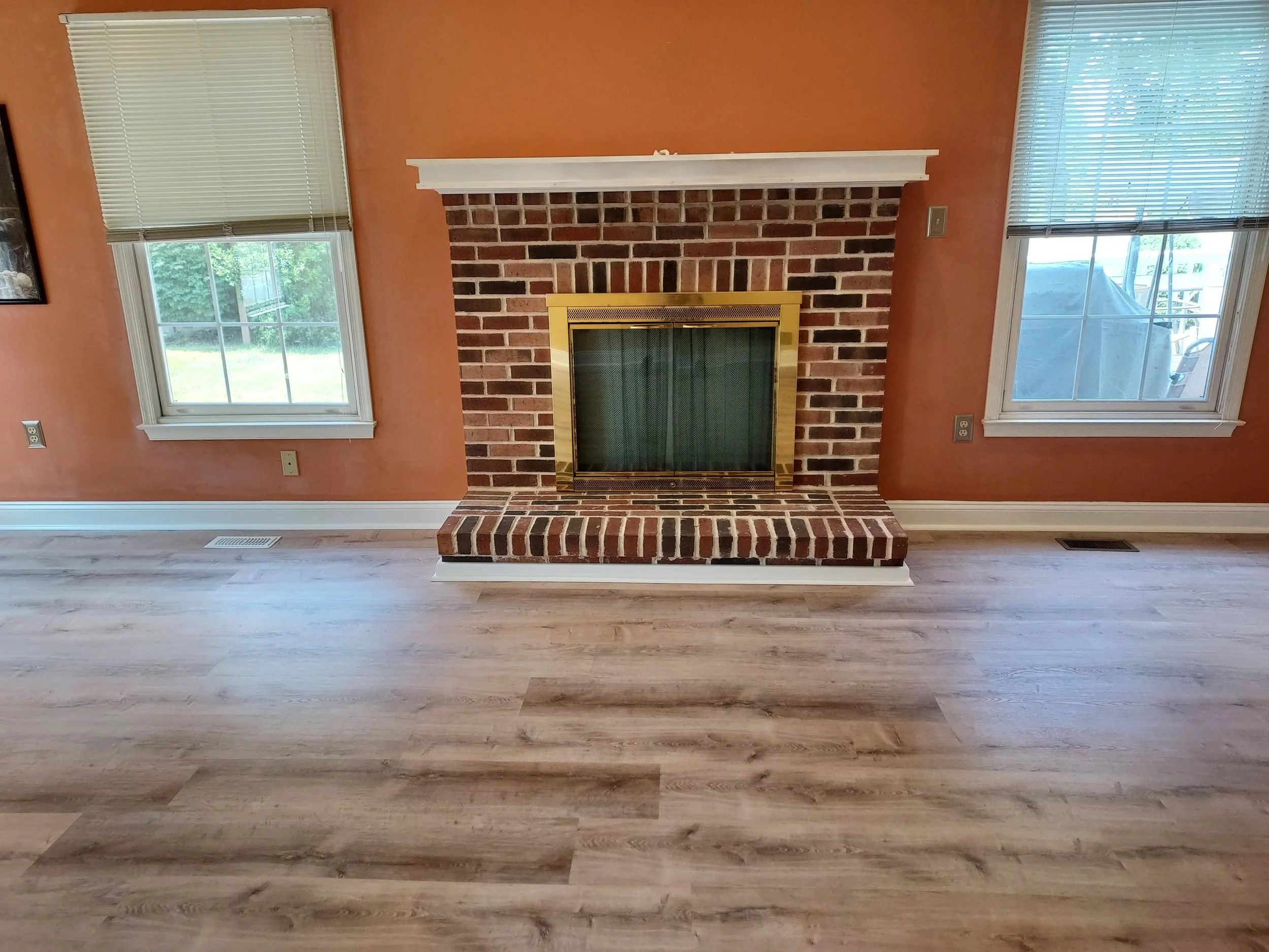 Living room with brick fireplace, two windows with blinds, orange walls, wood flooring, and a heating vent on the floor.
