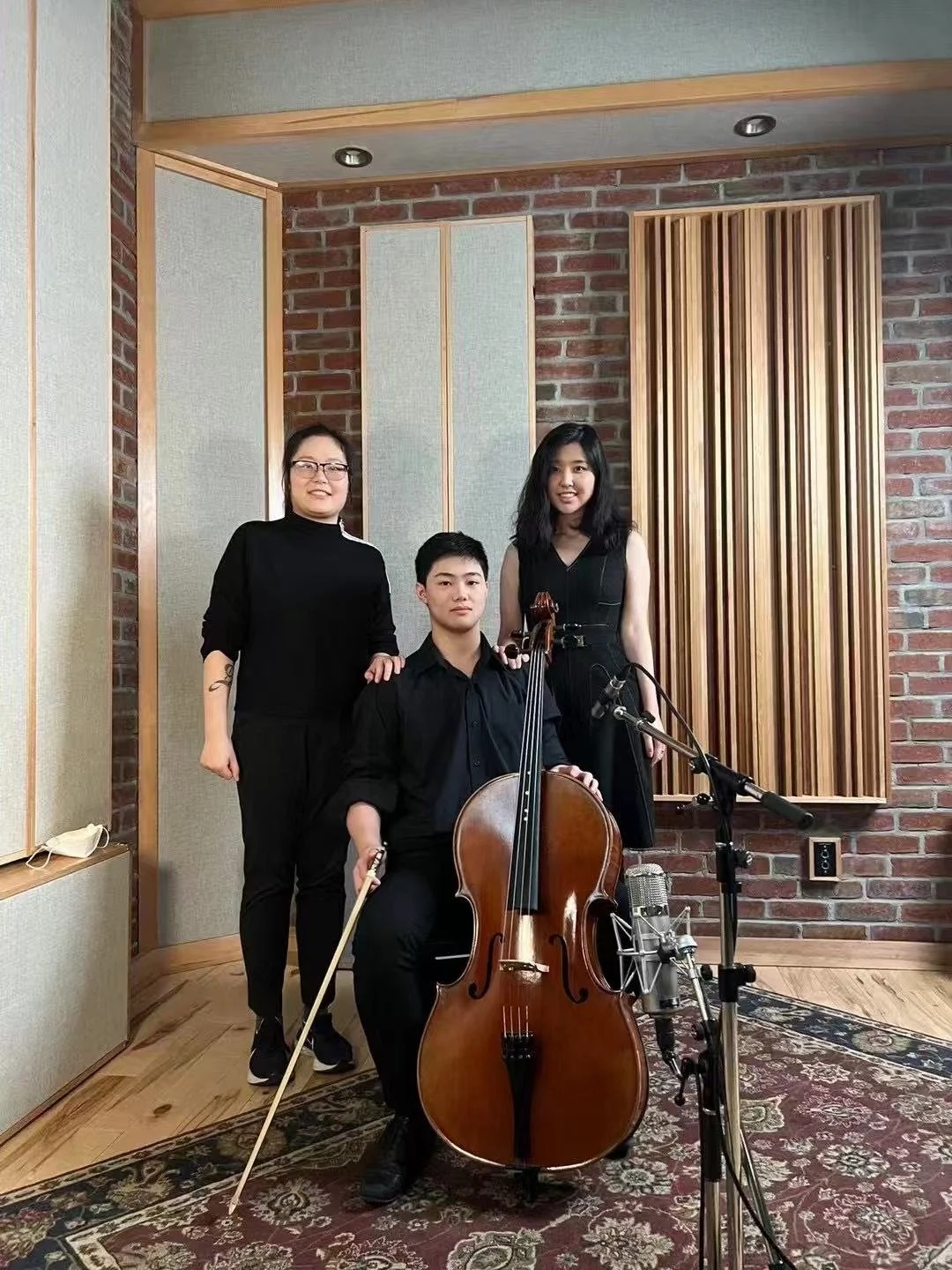 Three musicians in a recording studio with brick walls and acoustic panels. One is seated holding a cello and bow, with a microphone nearby. Two women stand behind him, one with glasses and tattoos, both smiling.