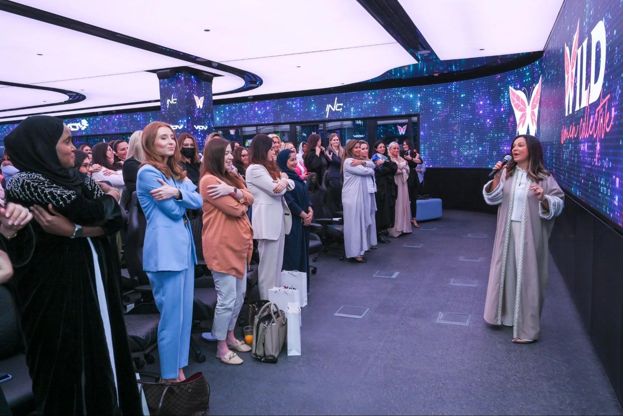 Women attending a presentation or conference listening to a speaker with a microphone, in a room with digital screens displaying a butterfly logo and digital graphics.