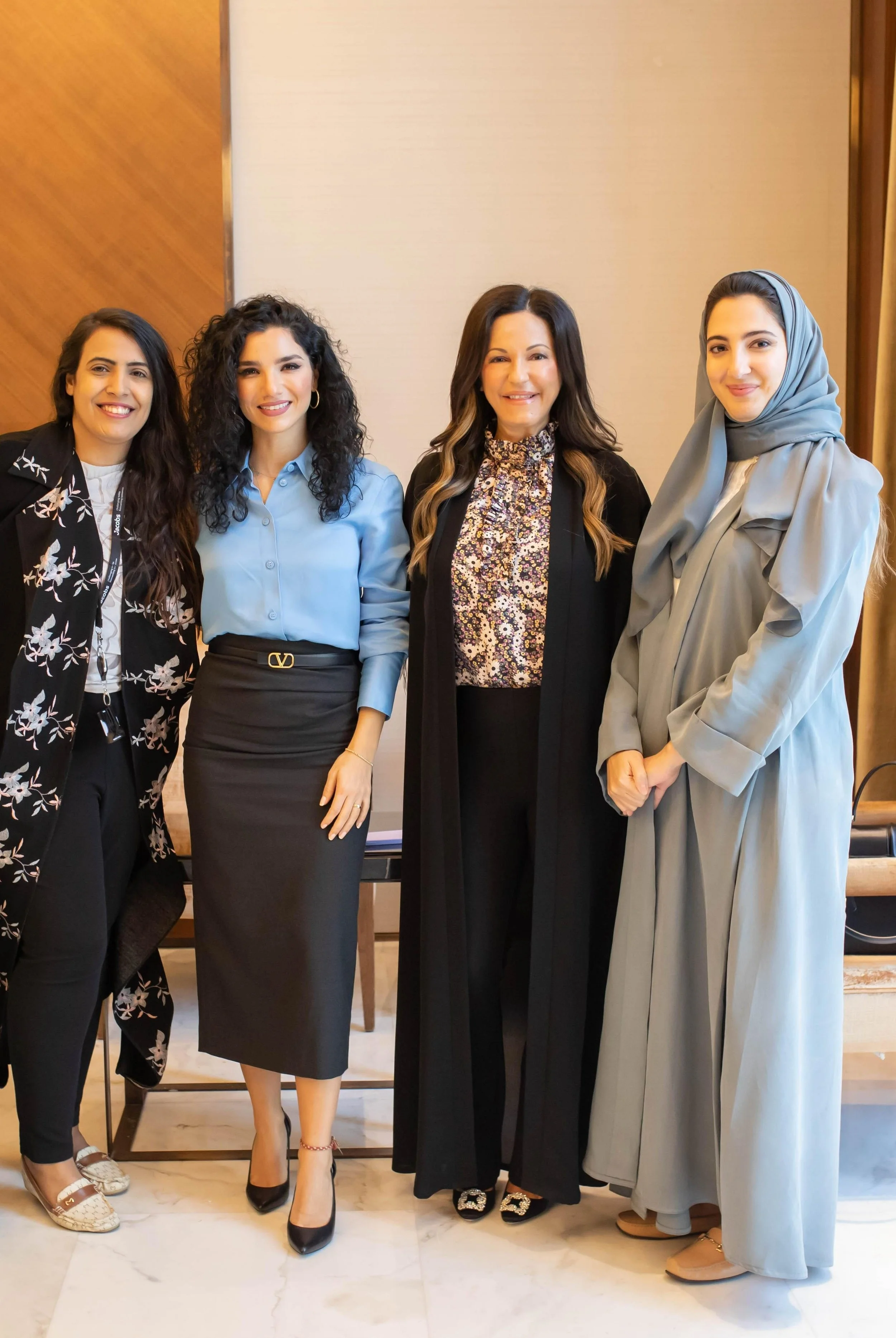 Four women standing together in a professional setting, dressed in business attire, with a wooden and beige background.