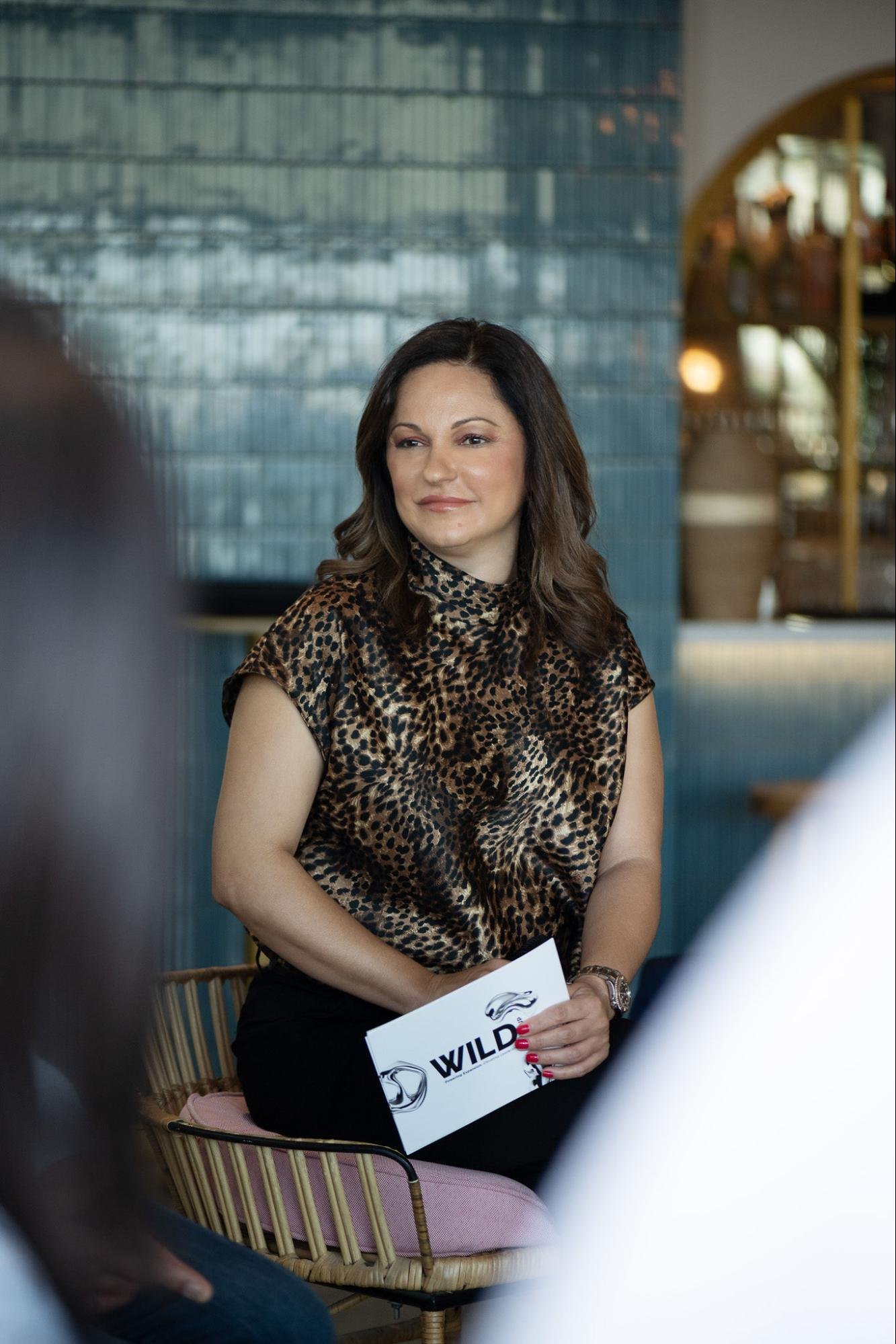 A woman with dark brown hair in waves, wearing a leopard print top, is sitting on a pink and gold cushioned chair and holding a paper with the word "WILD" on it. She is smiling slightly and looking to her right in a relaxed setting with a blue tiled wall in the background.