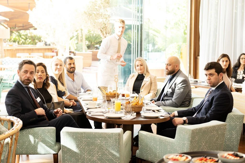 Business meeting with nine people seated around a round table in a bright restaurant. A waiter stands in the background.