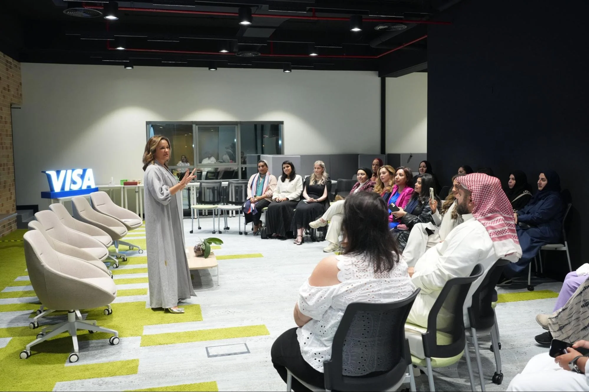 A woman in a gray coat is speaking or presenting in front of a seated audience, with a blue illuminated 'VISA' sign visible behind her. The audience includes women and men of diverse backgrounds, some women wearing hijabs, and one man in traditional Middle Eastern attire. The room is modern with black and white walls, bright lighting, and green carpet accents.